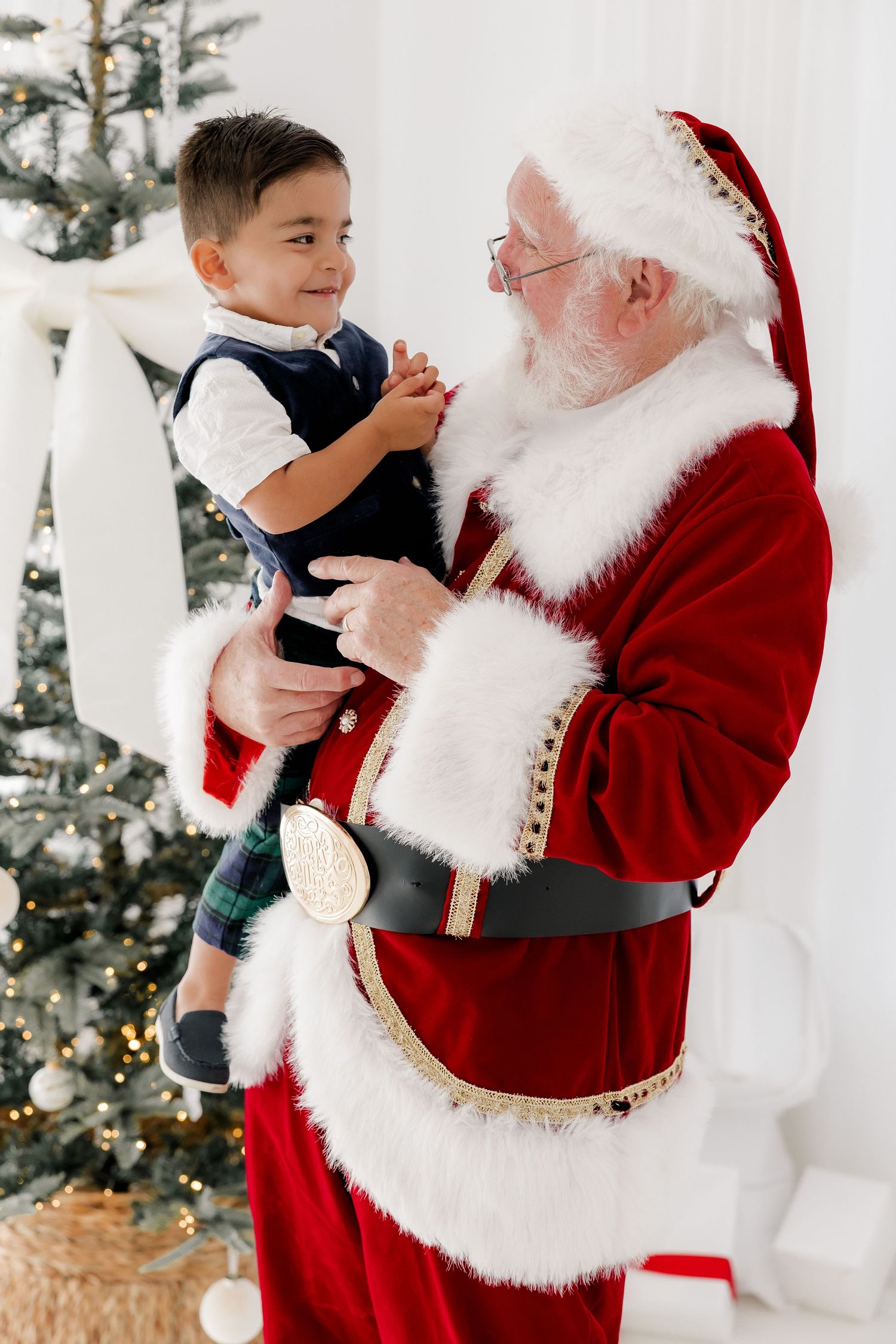 Santa Claus holds a smiling young boy near a decorated Christmas tree in a festive holiday portrait.
