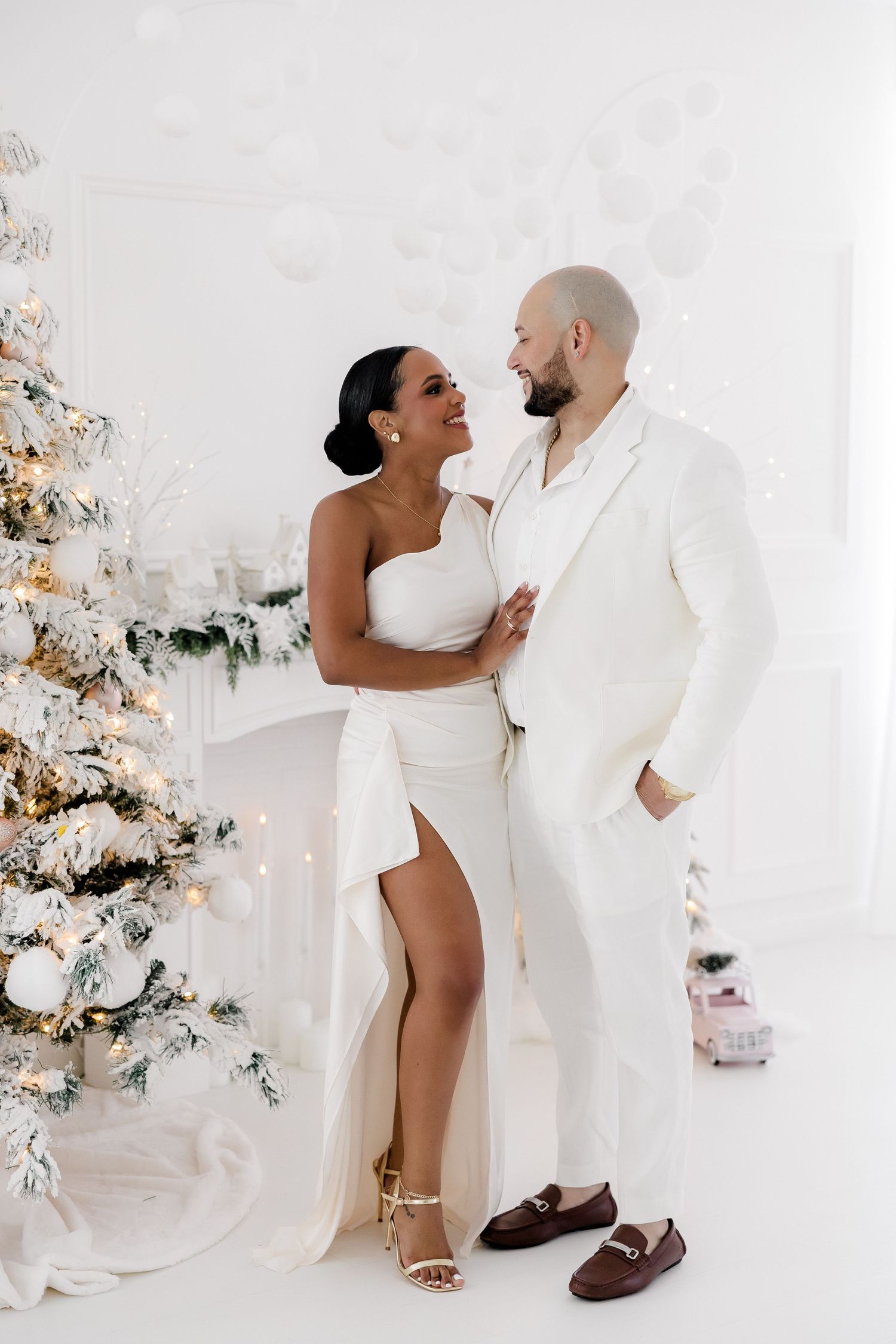 Romantic holiday photoshoot with couple in white outfits against decorated Christmas backdrop.