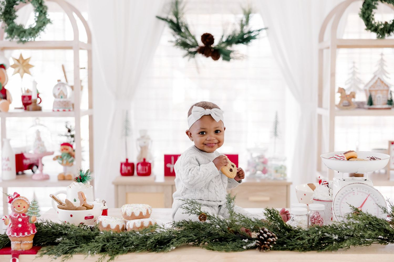 Baby sitting on white table with Christmas decorations and holiday displays in background.