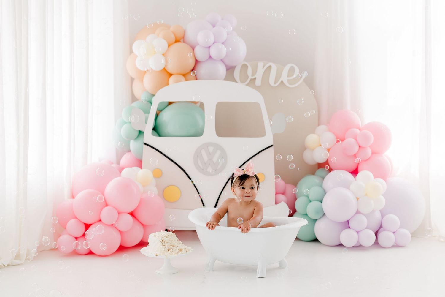 Baby girl sits in a white tub beside a VW bus prop, surrounded by pastel balloons for a first birthday cake smash.
