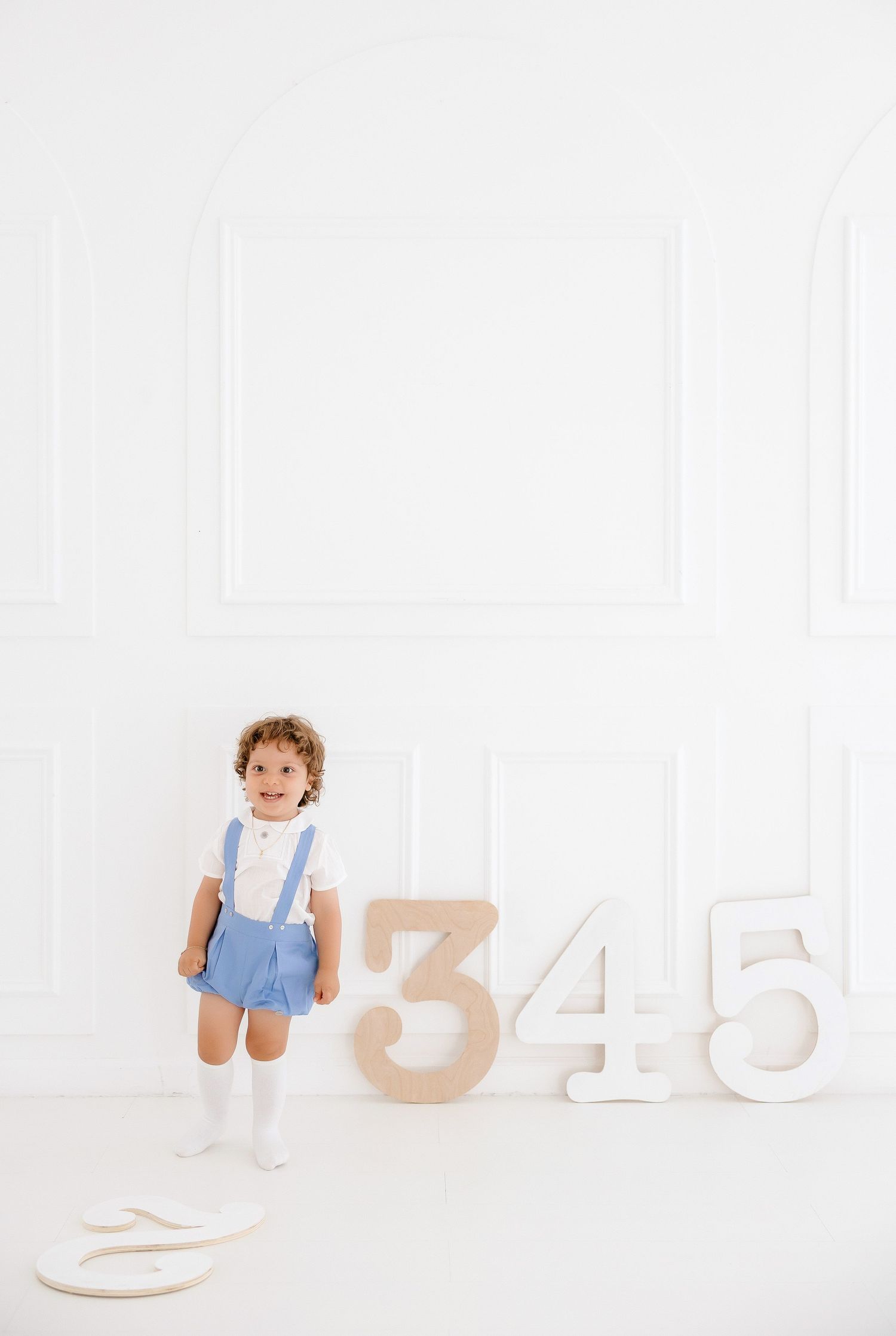 A baby in blue overalls sits among large white wooden numbers and letters against a bright white background.