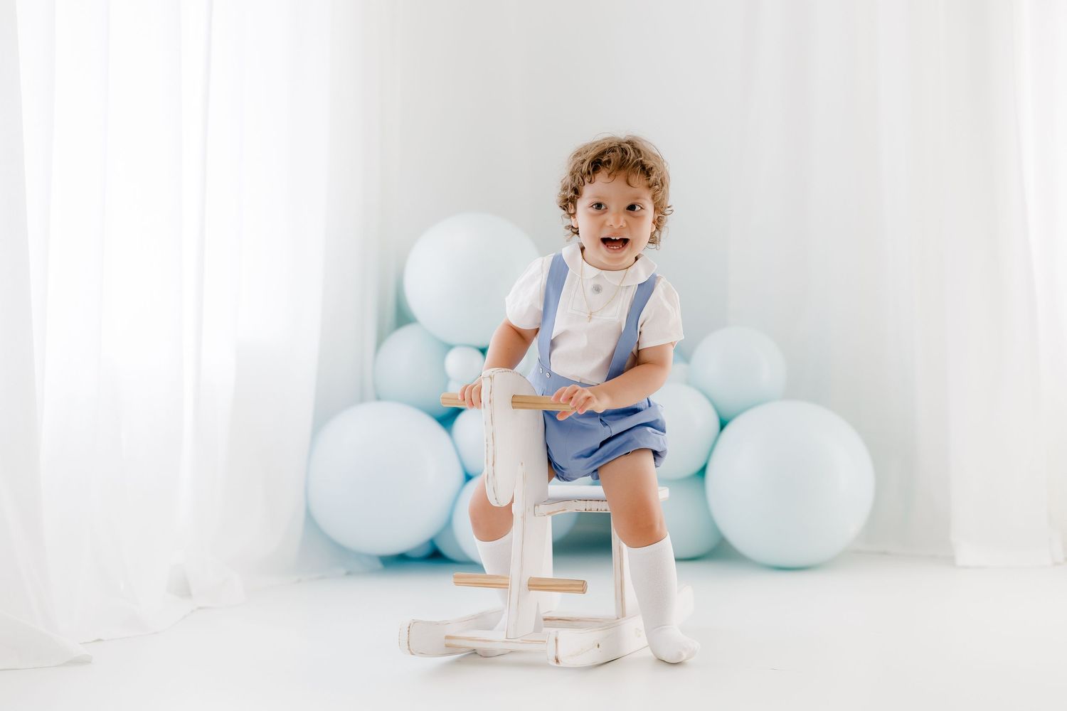 Baby in white outfit plays and smiles surrounded by soft blue balloons in bright studio setting.