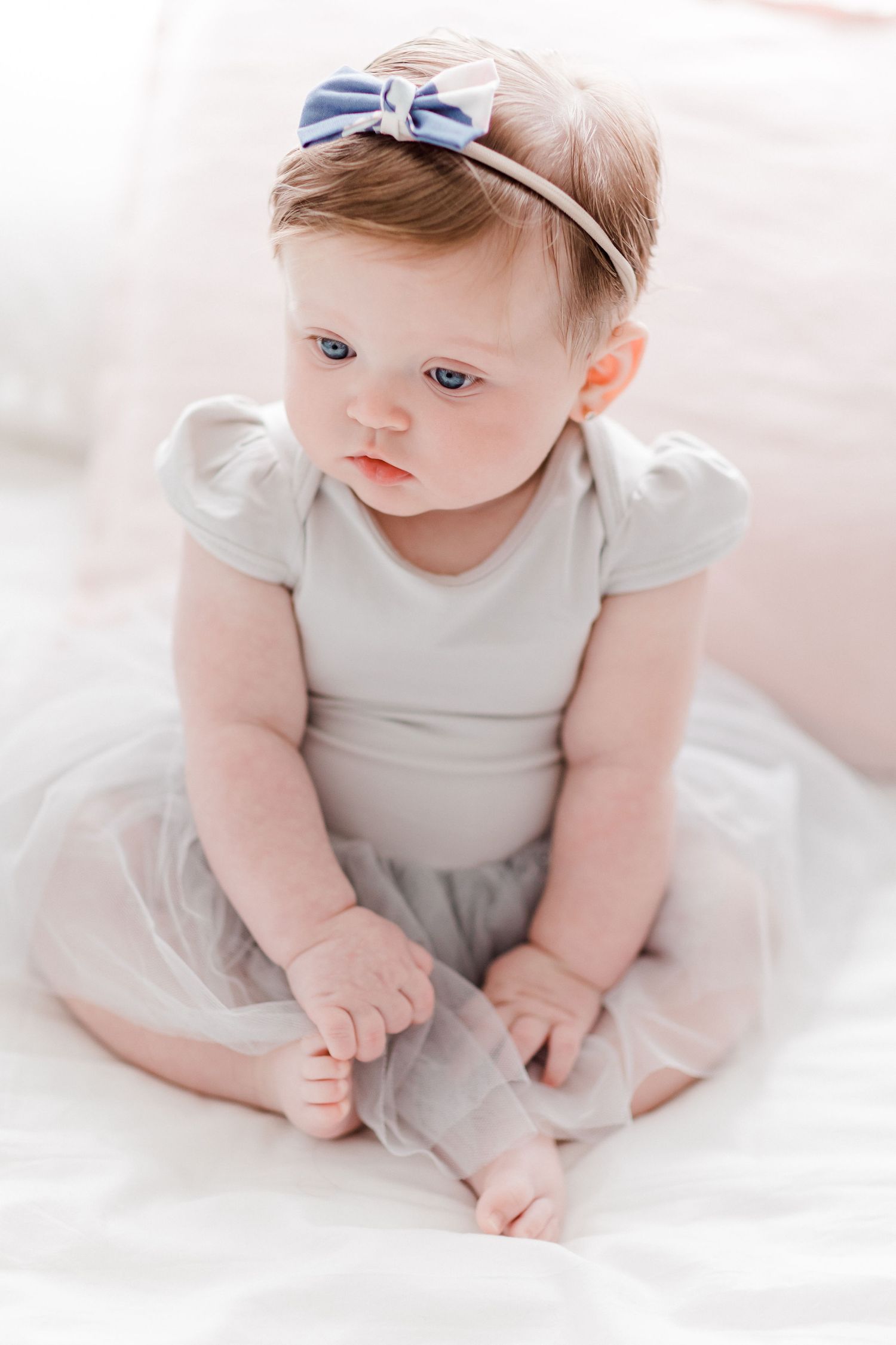 Sweet baby in white onesie with blue bow headband laying on soft white bedding.