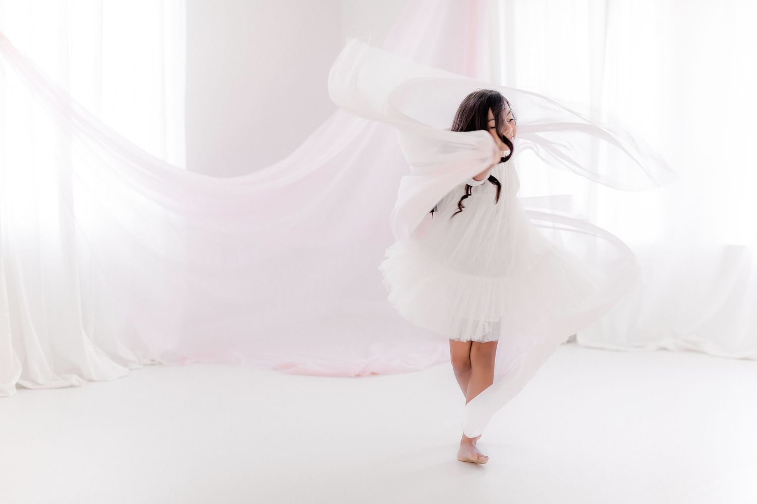A bride in a flowing white wedding gown poses elegantly against a light pink background.