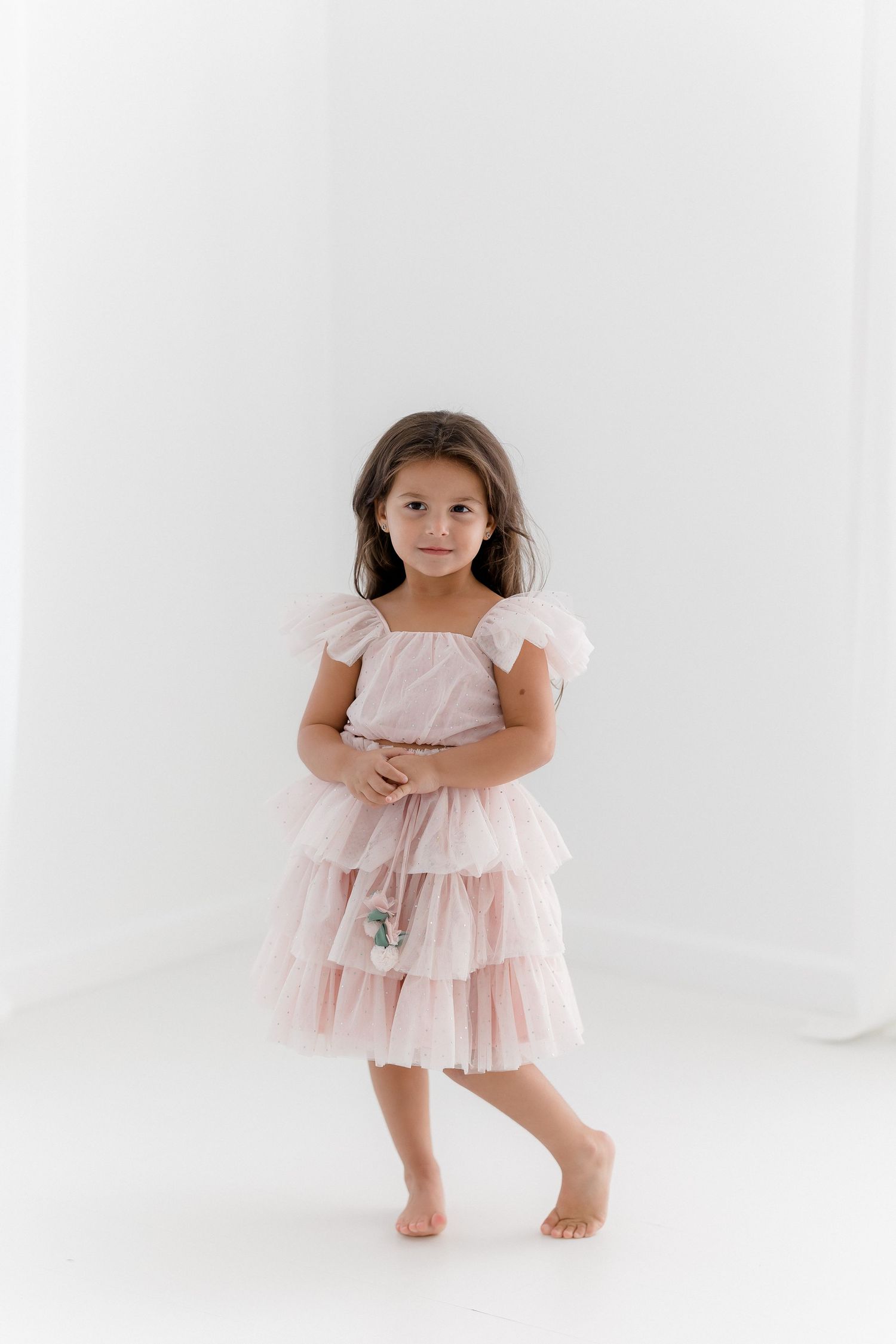 A young child wearing a pink tiered tulle dress stands barefoot against a white background.