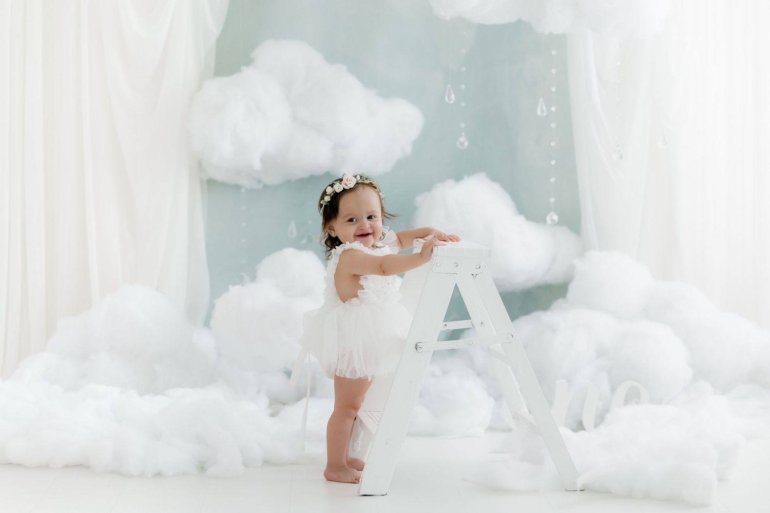 Adorable baby girl in white tutu dress standing among fluffy clouds in a whimsical dreamy studio photo session.