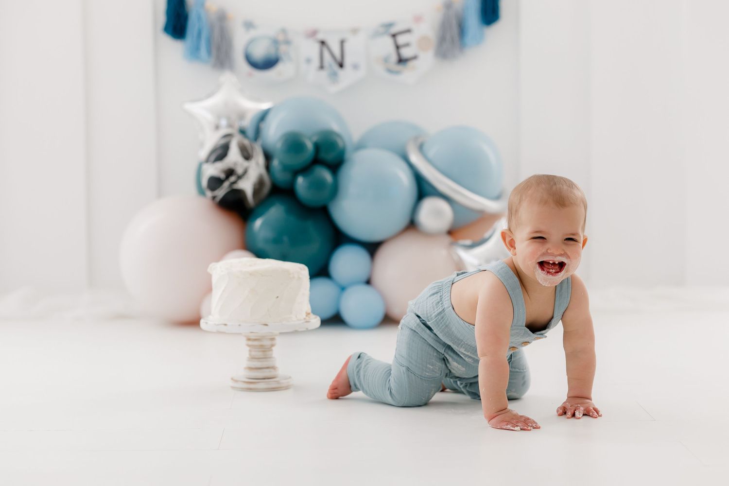 First birthday setup with blue and white balloons and cake against white backdrop.