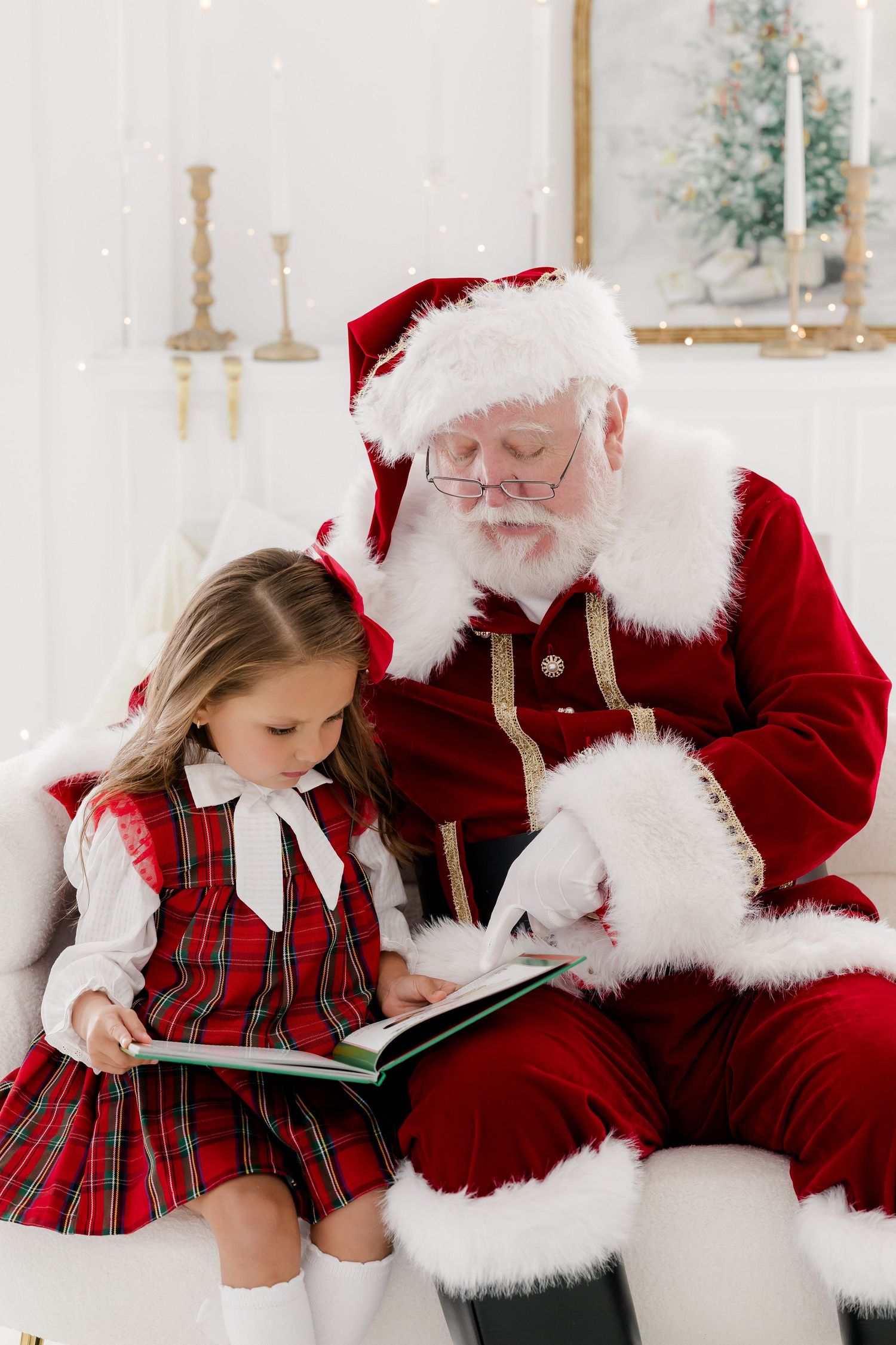 Santa Claus reads a Christmas book with a young girl in a plaid dress in a festive white holiday setting.