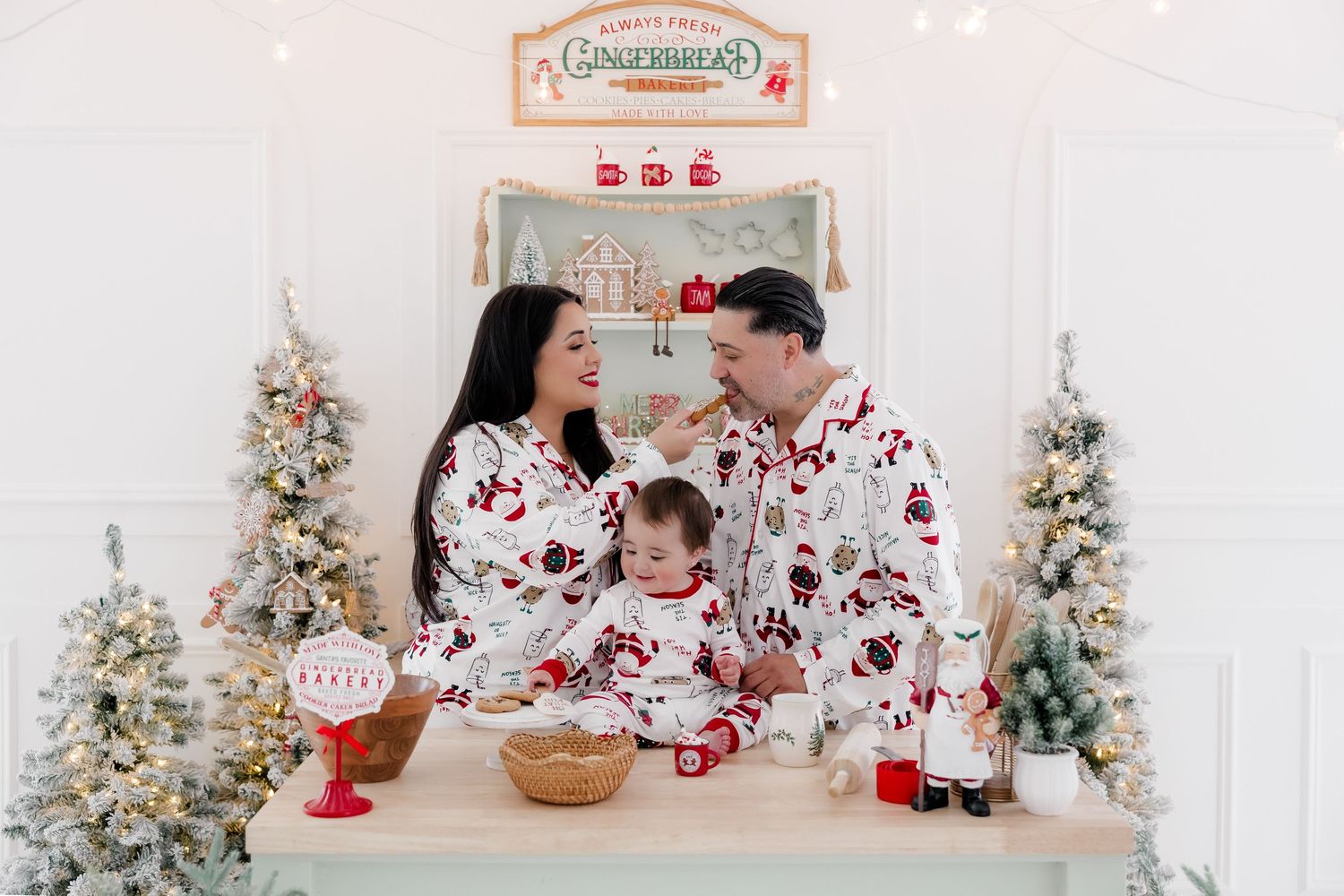 Family in matching Christmas pajamas posing at a gingerbread bakery themed holiday photo set with festive trees.