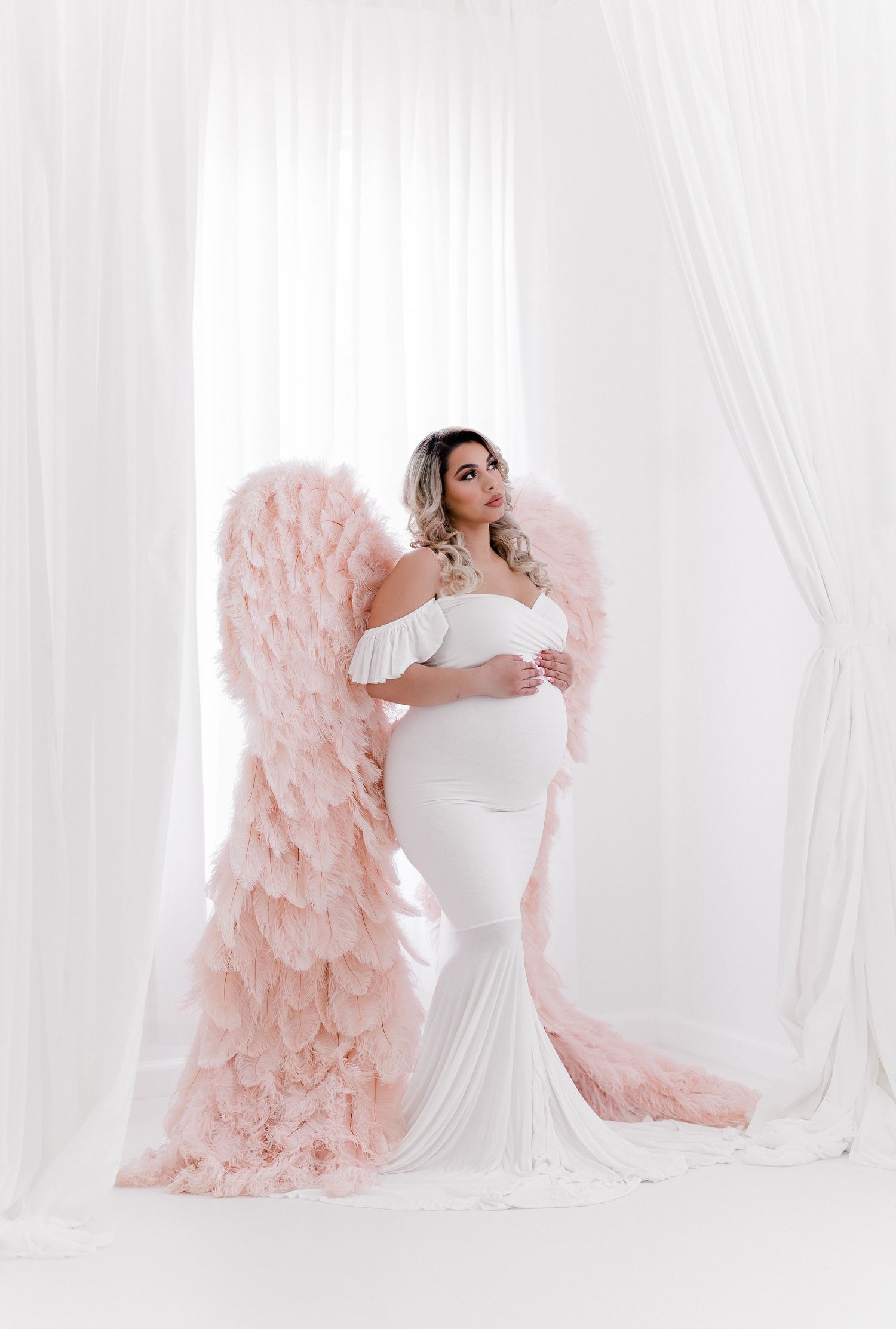 Romantic maternity photos with pink ruffled chair and white gown in bright studio setting.