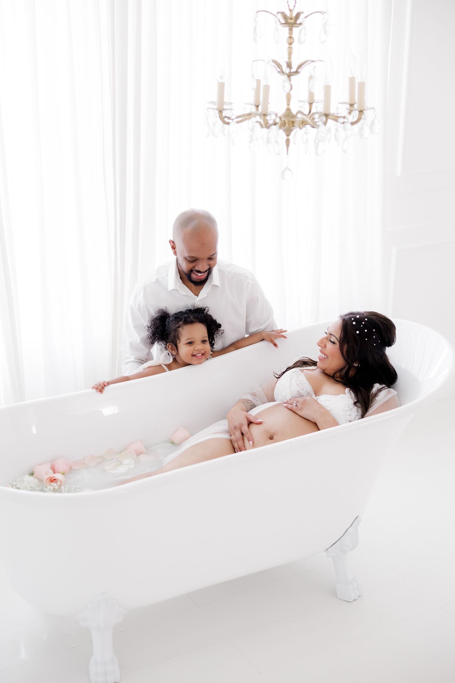A serene milk bath photography session shows a family sharing intimate moments in an elegant white bathroom setting.