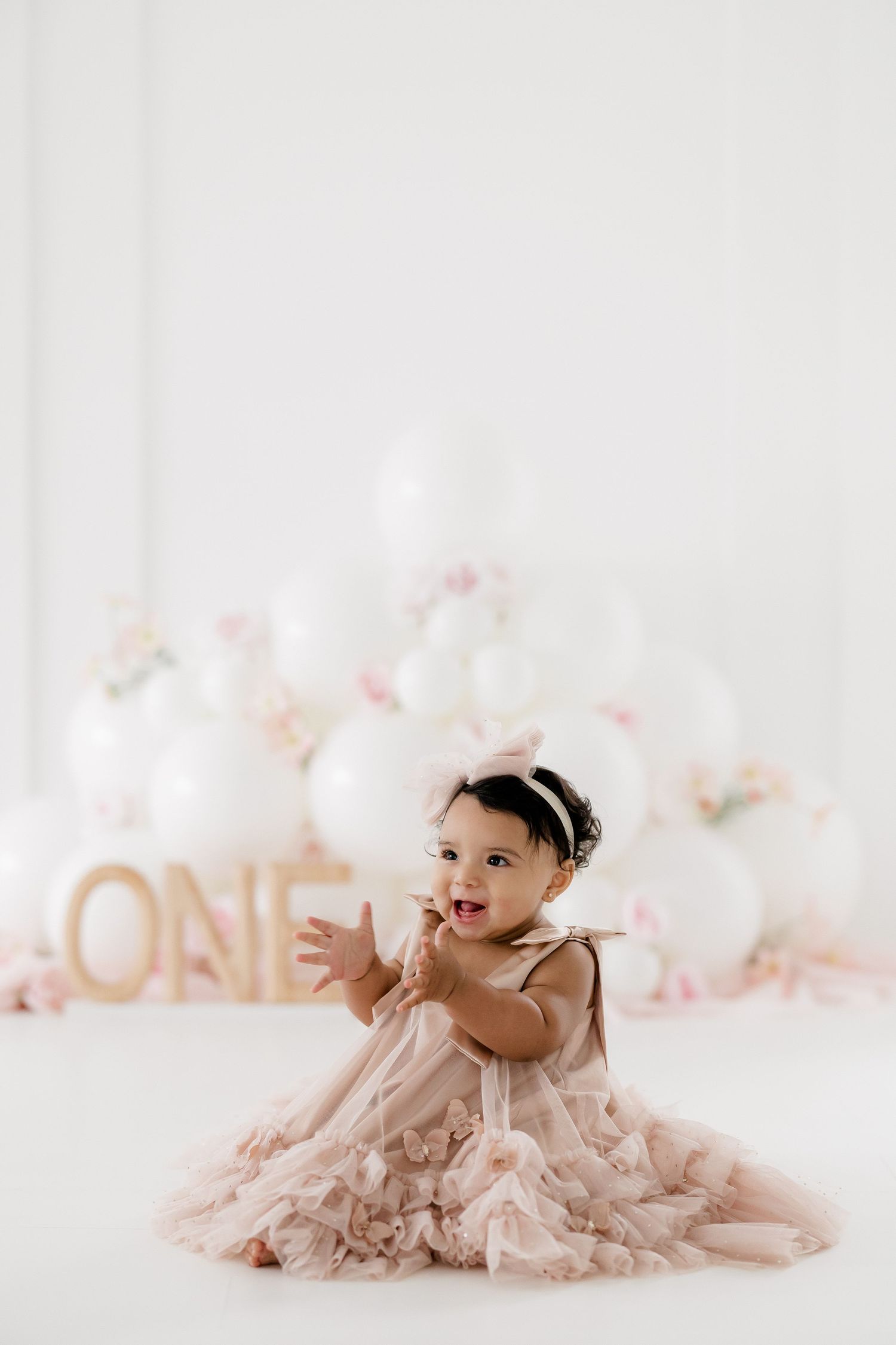 A baby in a pink tulle dress sits on white floor with balloon decorations and gold ONE letters behind them.