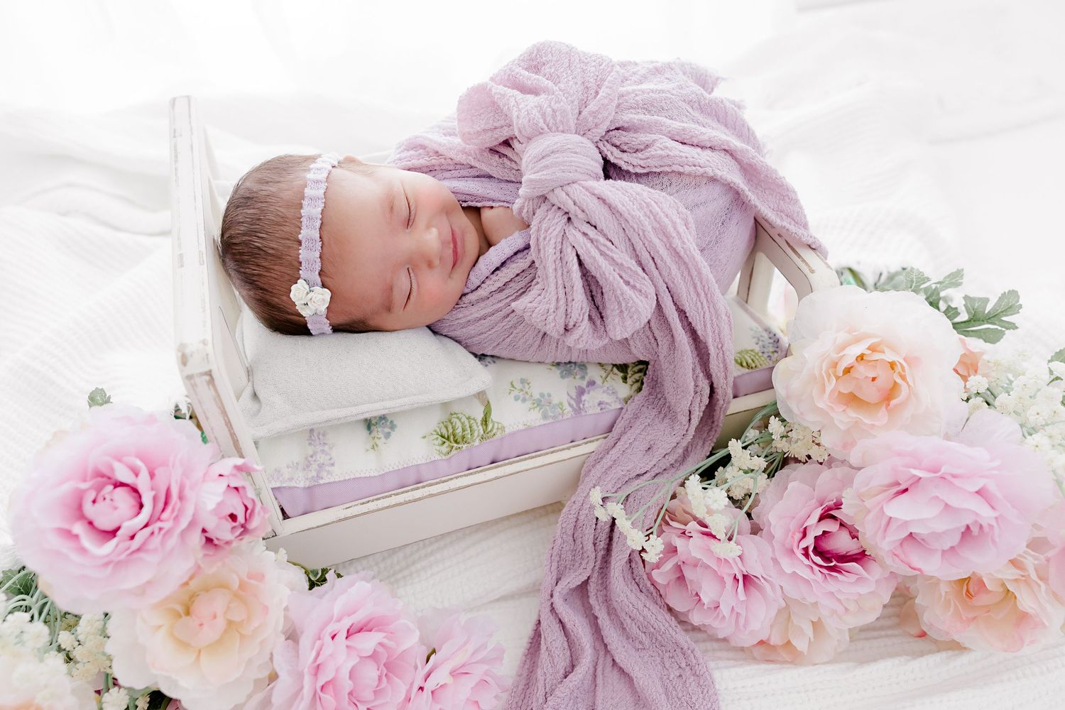 Artistic newborn portrait with pink roses and flowing purple fabric in minimalist style.