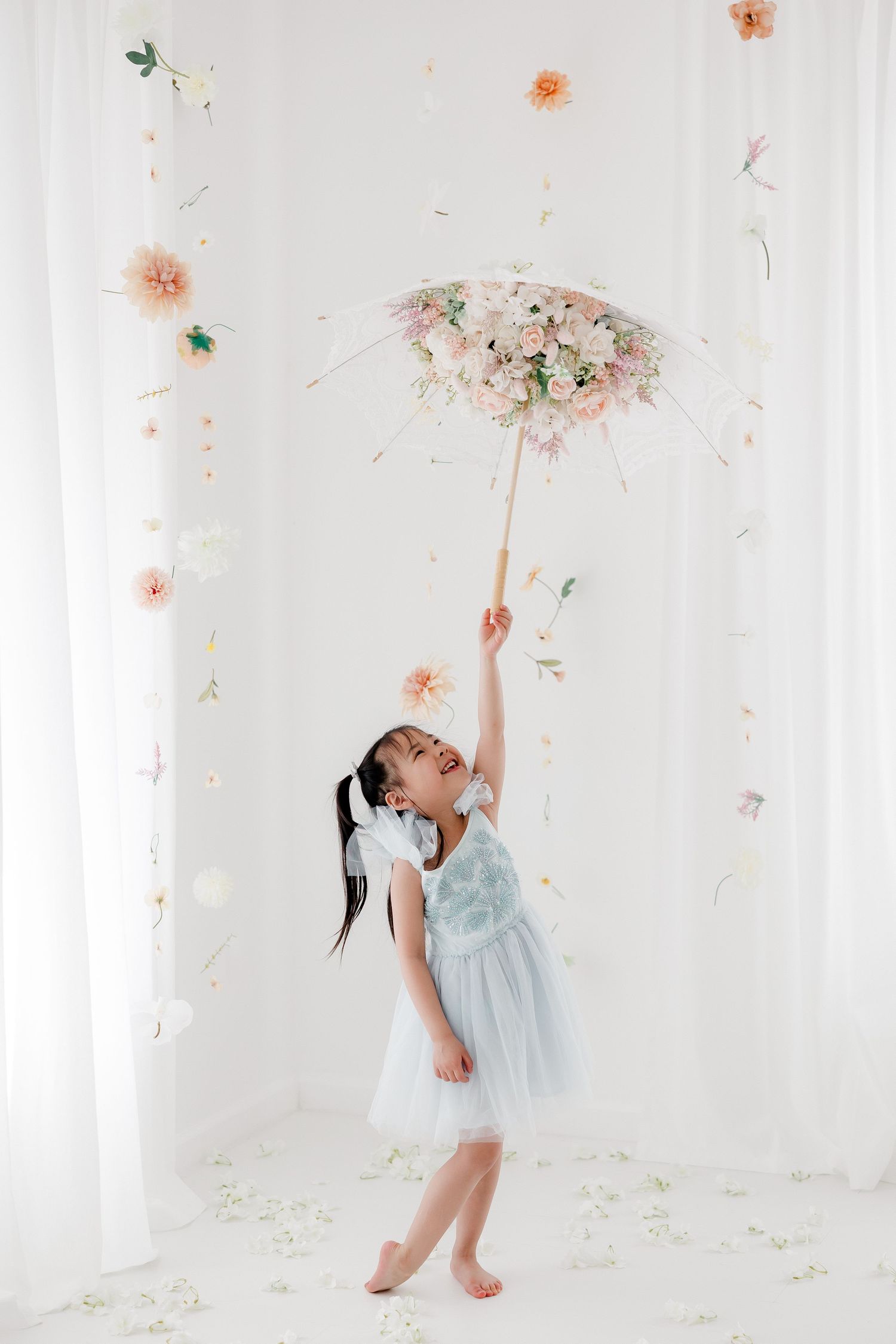 A dancer in a light blue dress twirls gracefully among falling pink flower petals in a bright white studio setting.
