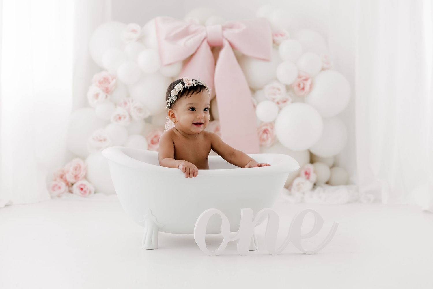 A baby sits in a white bathtub surrounded by soft pink flowers and decorative elements in a dreamy studio setting.