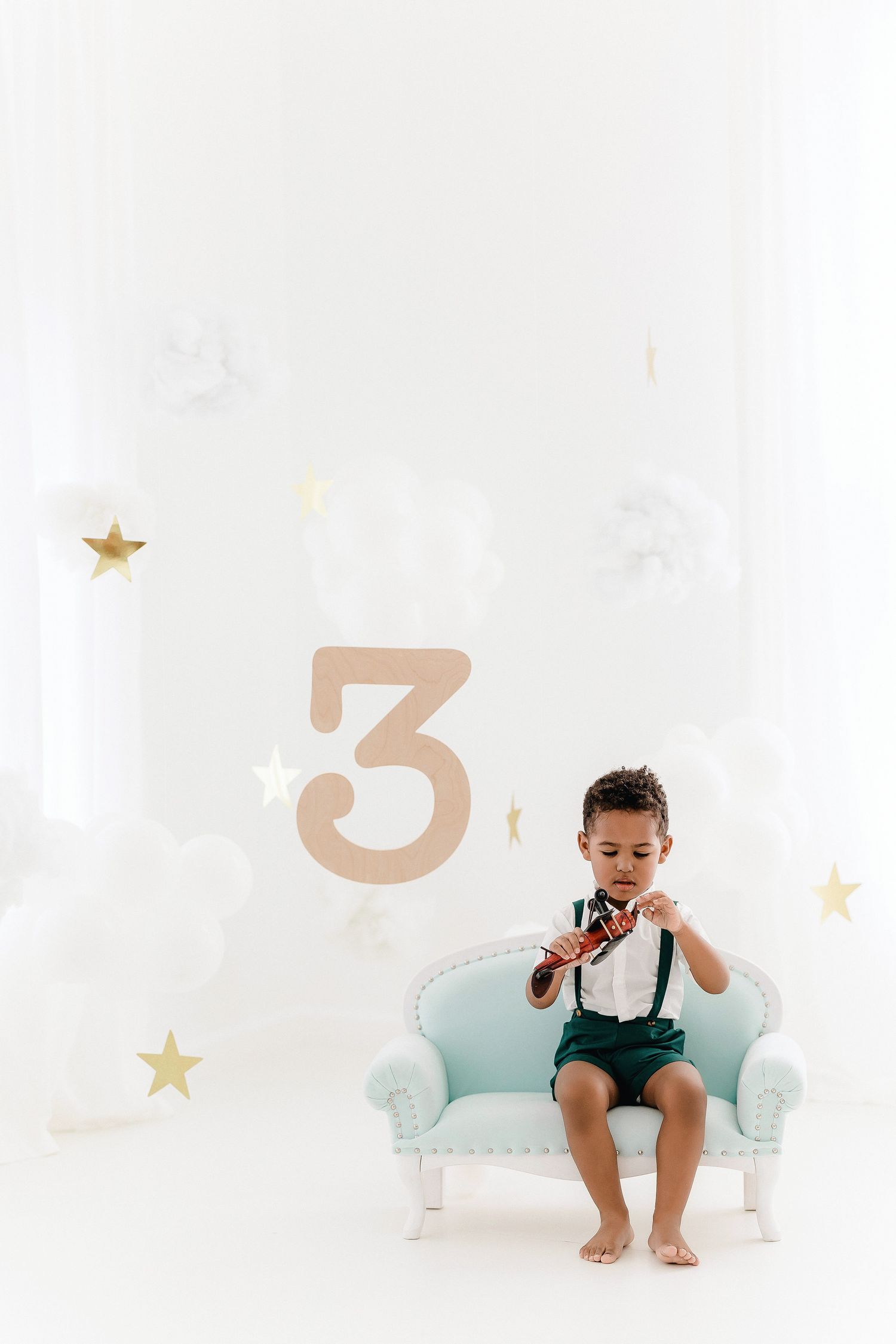 Baby in turquoise shorts sits on white couch with gold star decorations in dreamy nursery setting.