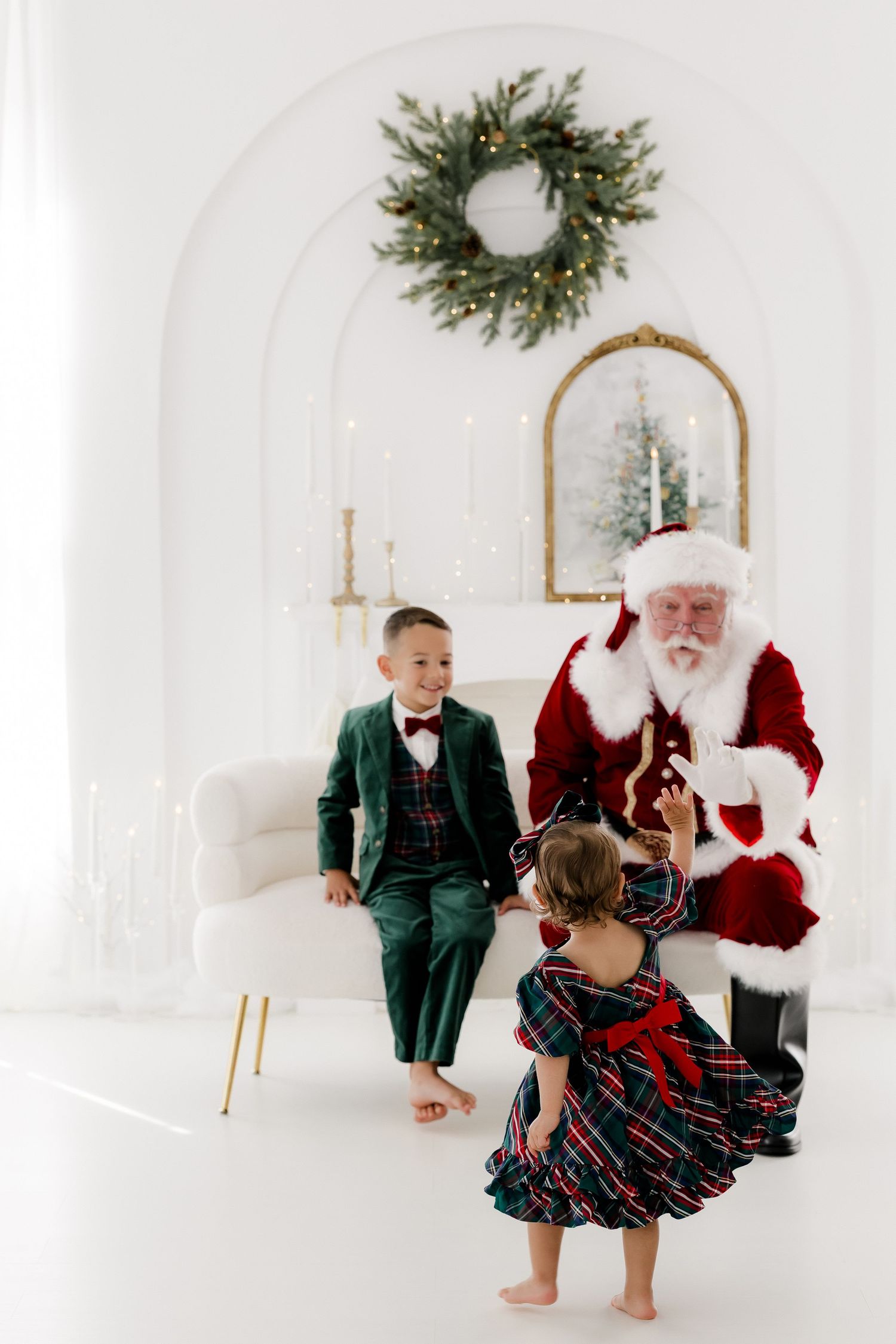 Children in festive attire meet Santa Claus in a bright white Christmas decorated room with wreath and mirror.