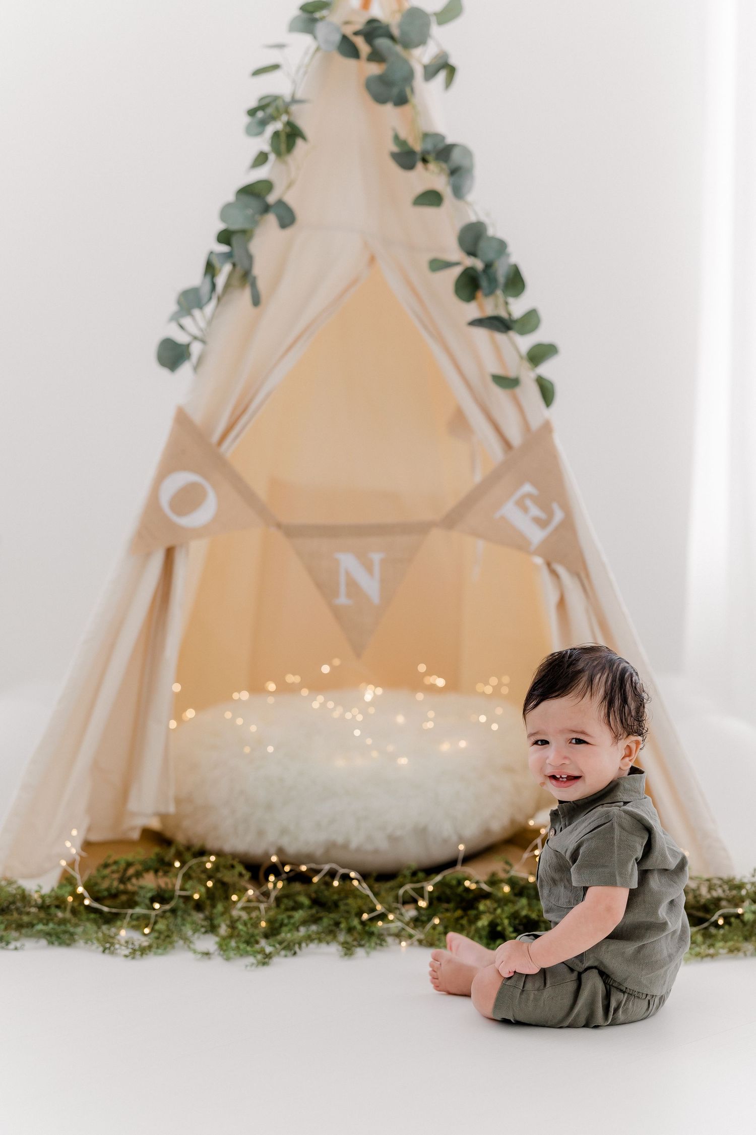 A small child in olive green clothing sits near a white teepee tent decorated with fairy lights and greenery.