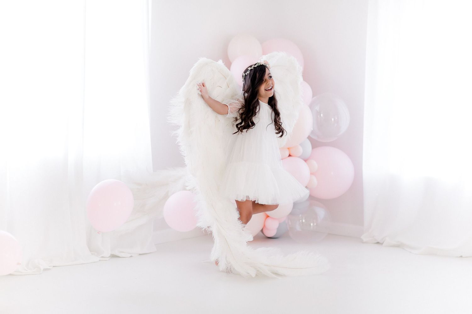 Ethereal white dress and feathers create dreamy scene with soft pink balloons in minimalist white studio setting.