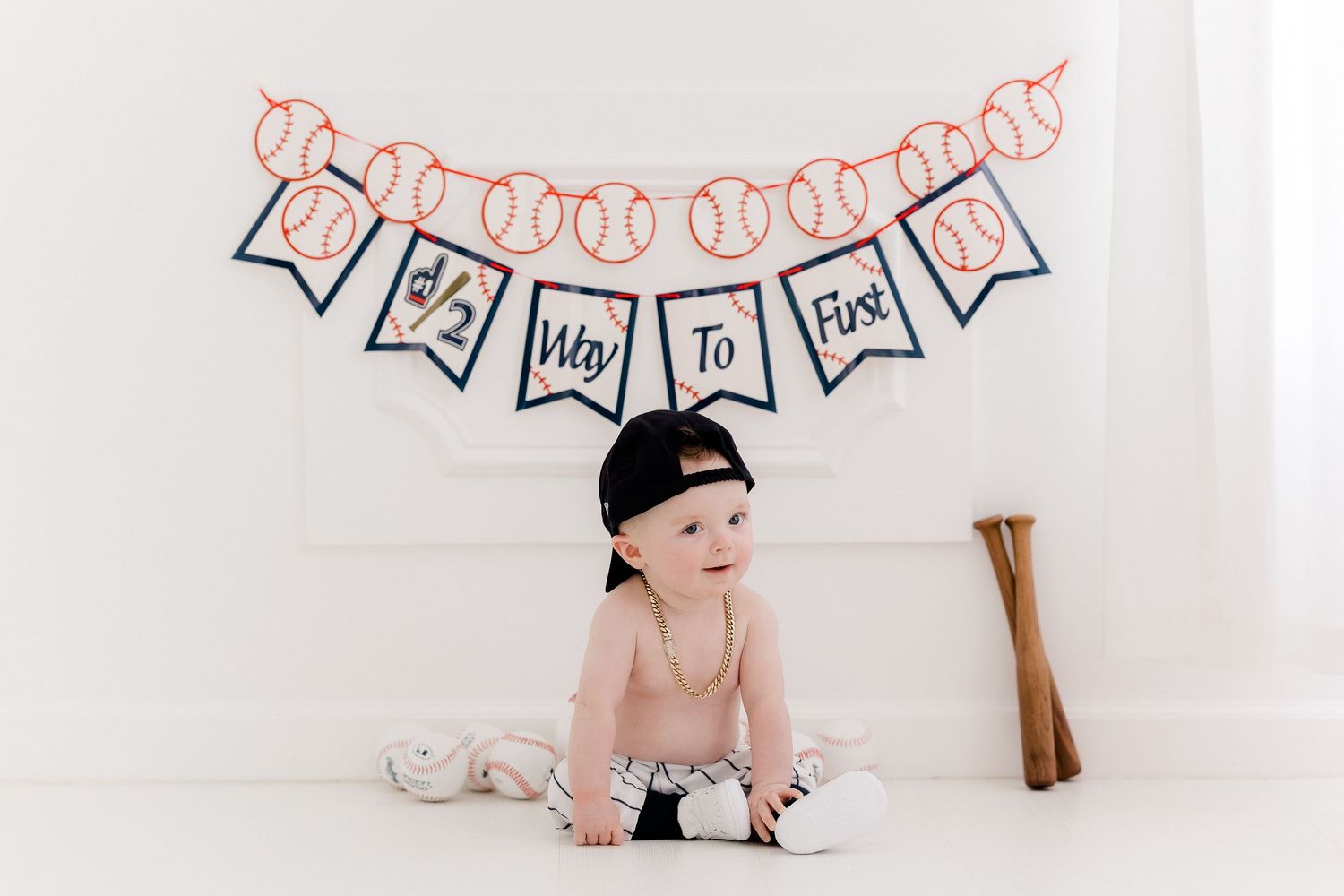 Baby wearing black baseball cap sits beneath decorative banner during milestone photo session with baseball bat prop.