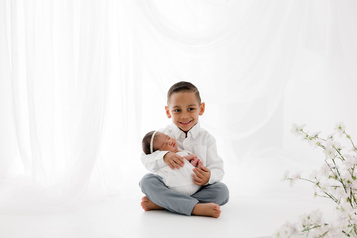 A child in a white shirt and jeans sits cross-legged holding a swaddled newborn against a bright white backdrop with flowers.