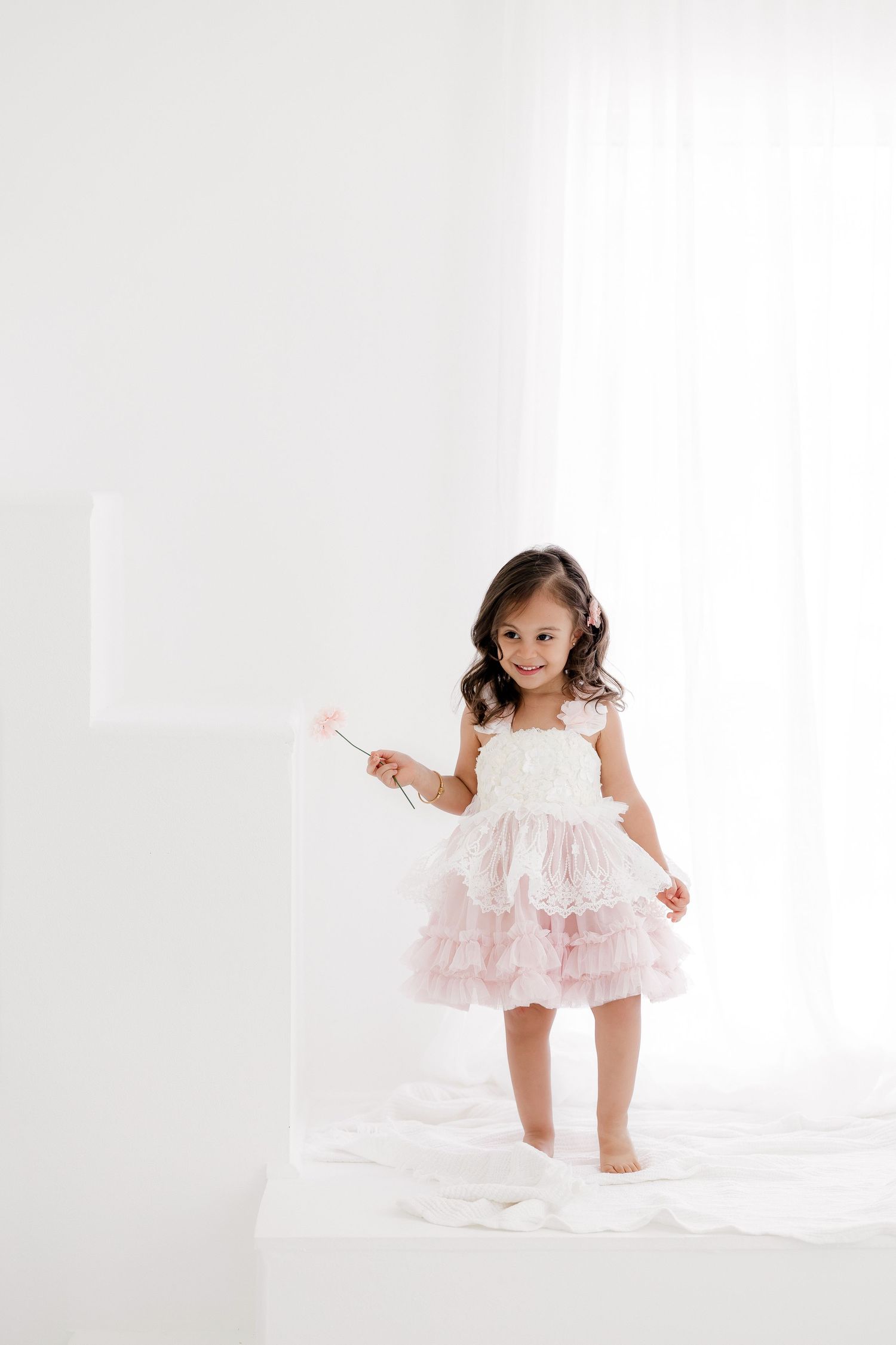 A young child in a white and pink ruffled dress twirls and plays in a bright white studio setting.