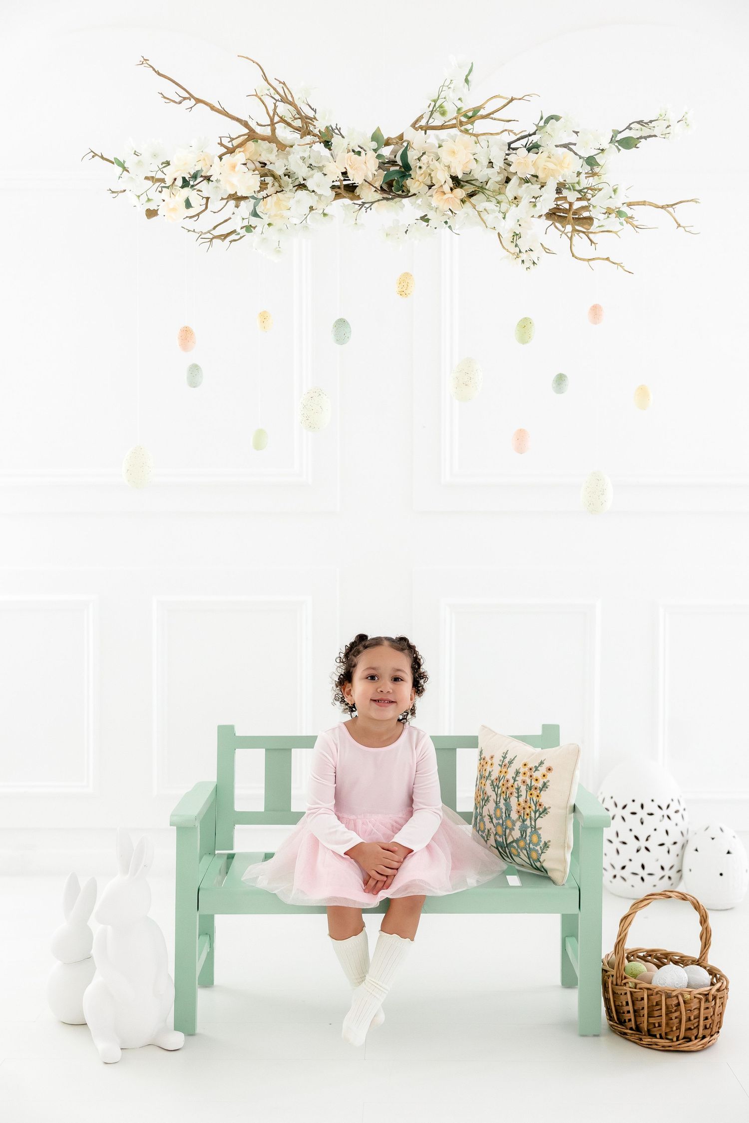 Baby in pink dress sits on mint green bench with basket and floral decorations in white studio setting.