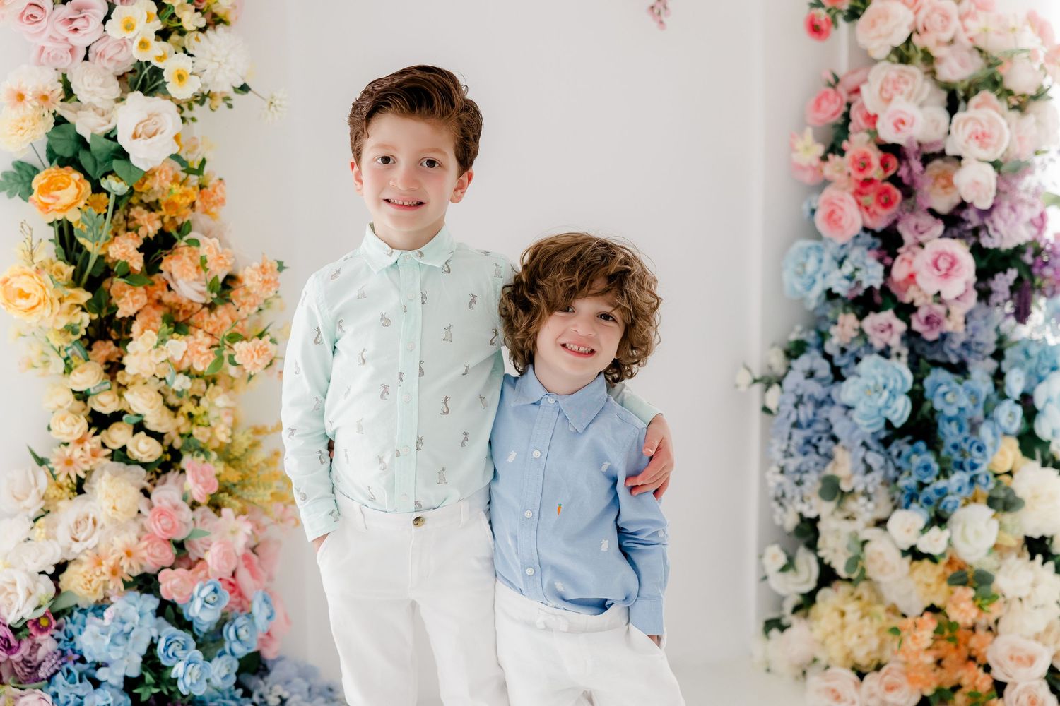 Two young children pose together against a backdrop of colorful floral arrangements in pastel blue, pink, and yellow.