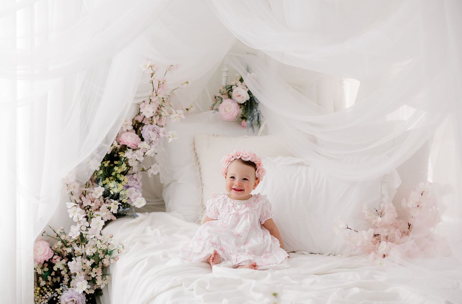 Baby in white dress and bonnet sits surrounded by delicate pink and white cherry blossoms in a dreamy studio setting.