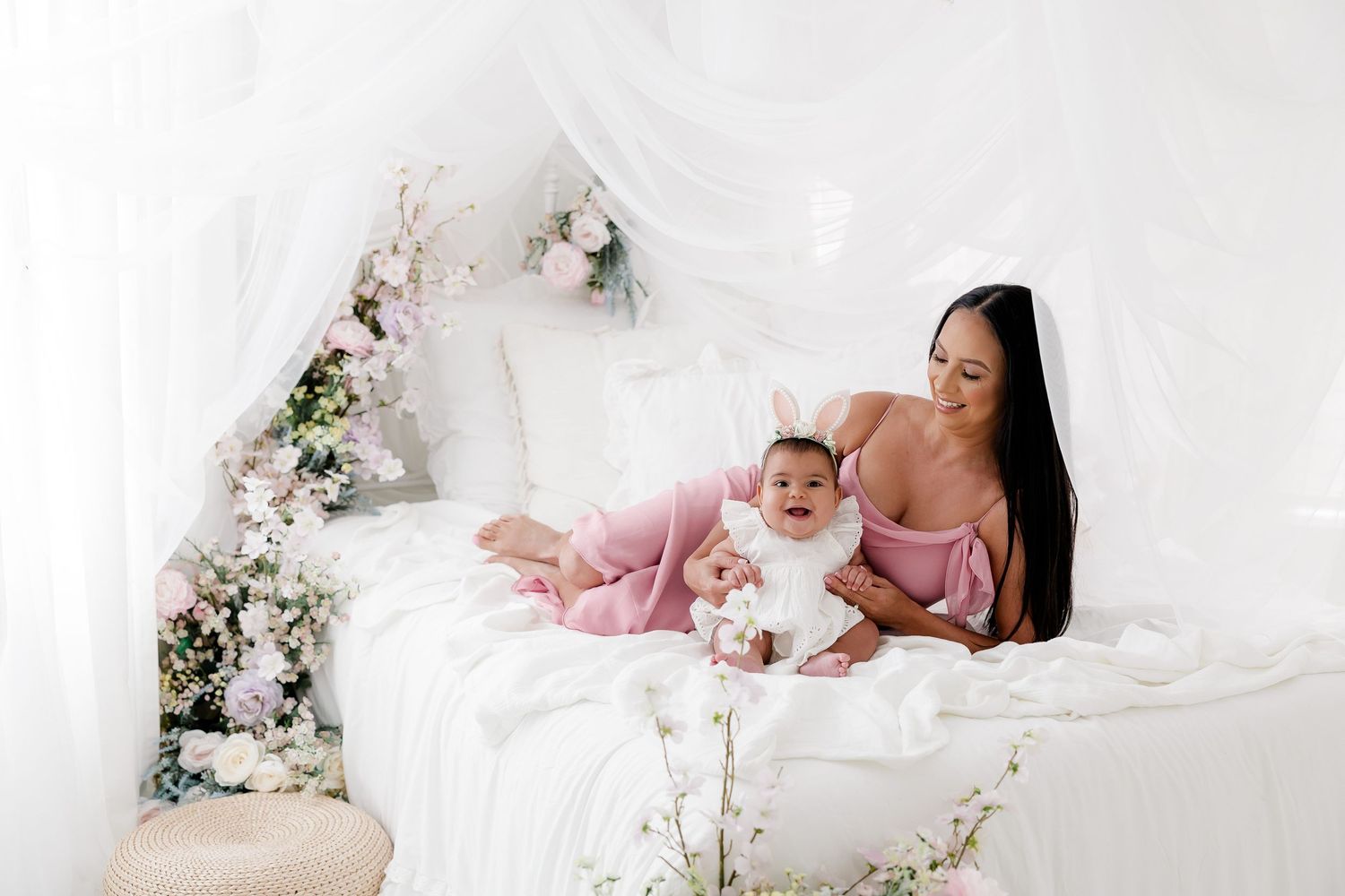 A newborn baby lies peacefully on white bedding surrounded by delicate pink and white floral arrangements.