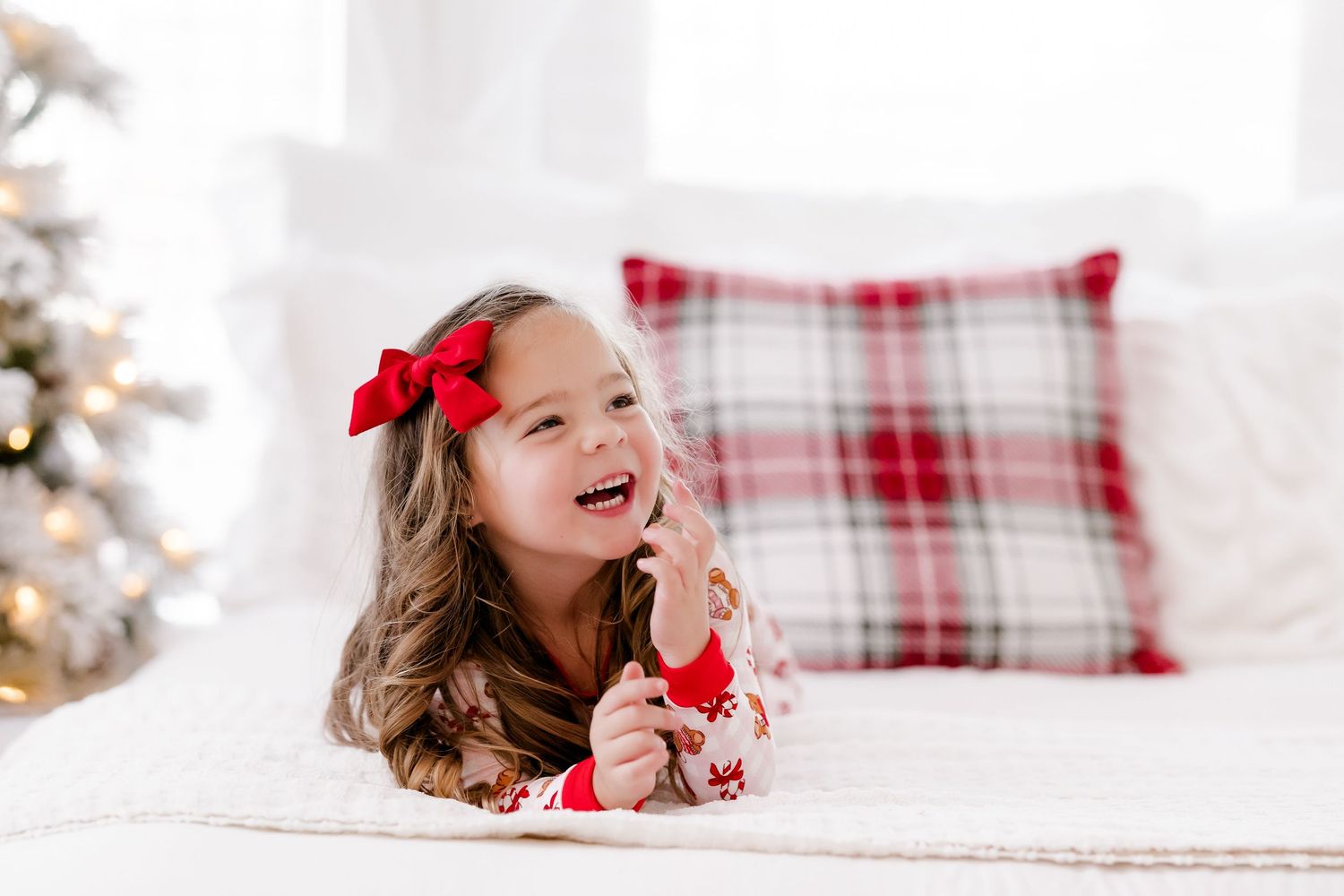 Young girl in Christmas pajamas laughing on white bed with plaid pillow and holiday tree in background.