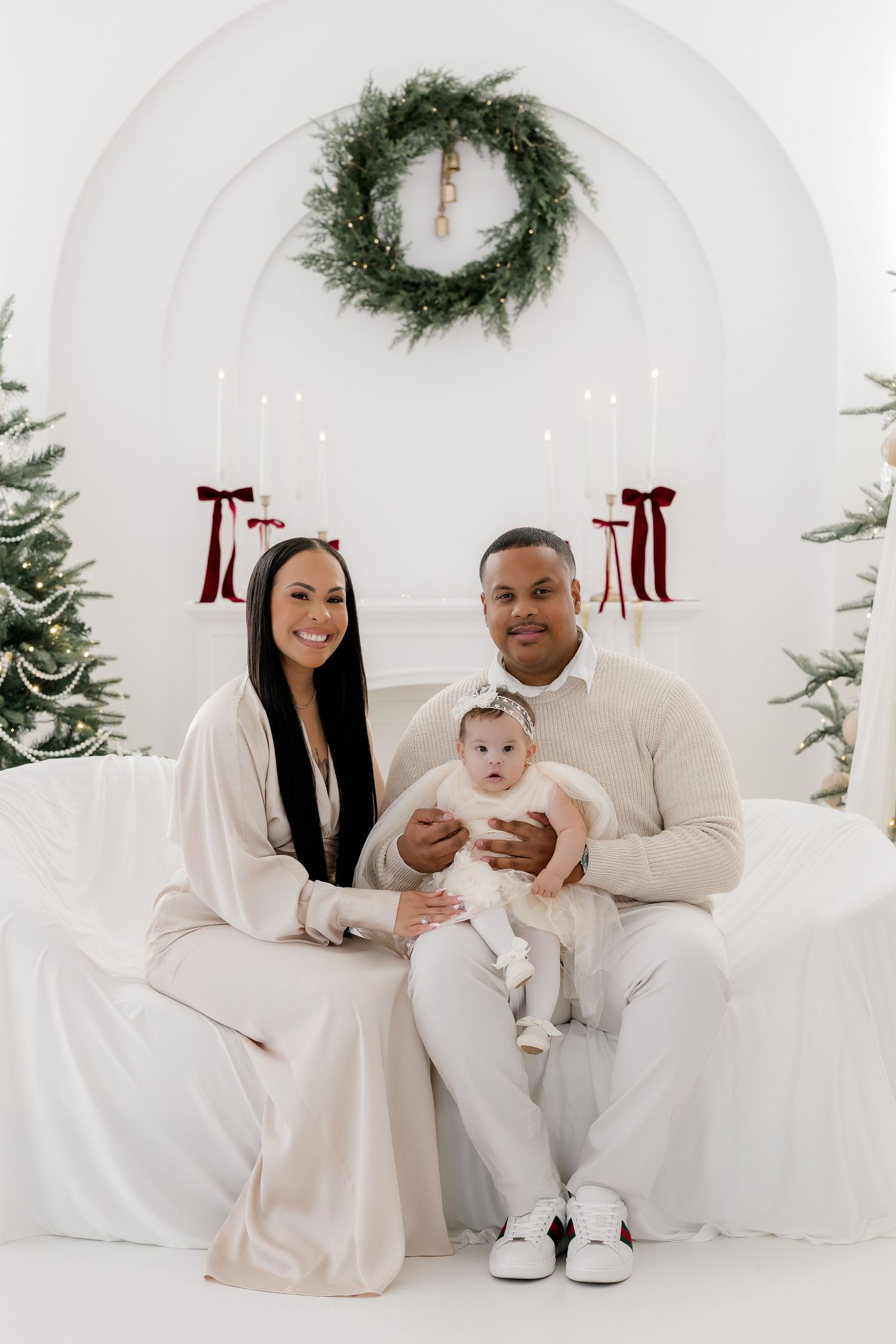 Family Christmas portrait with parents and baby in white outfits, seated before festive wreath and candle decor.