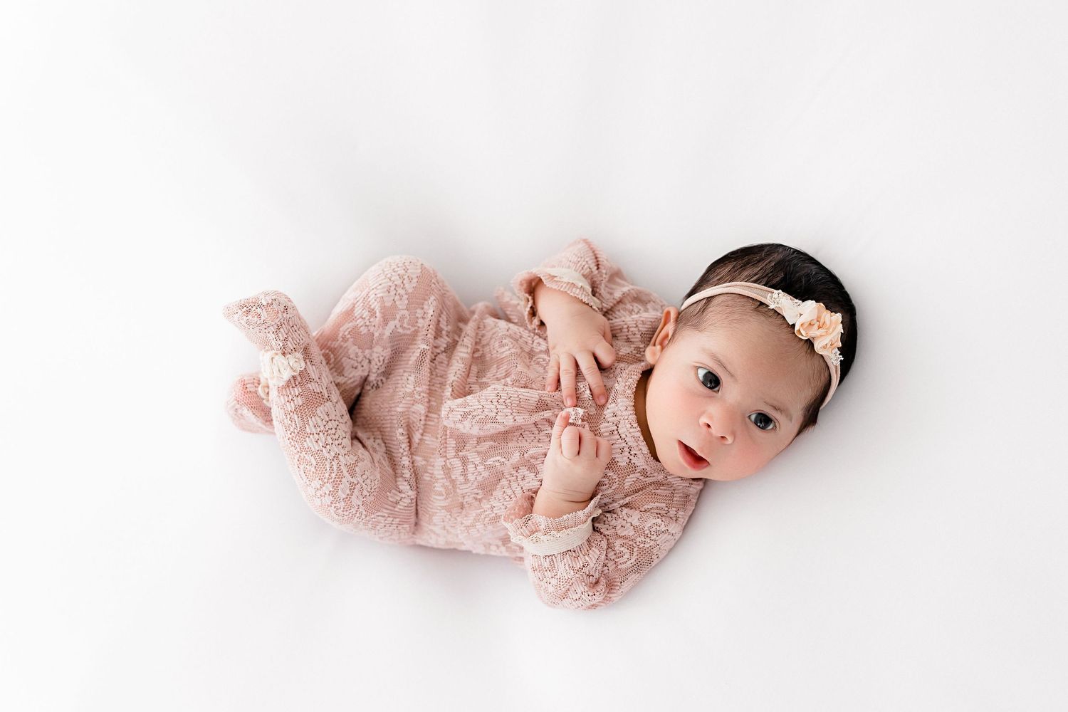 Baby in pink lace outfit with headband lying on white surface during newborn photoshoot.