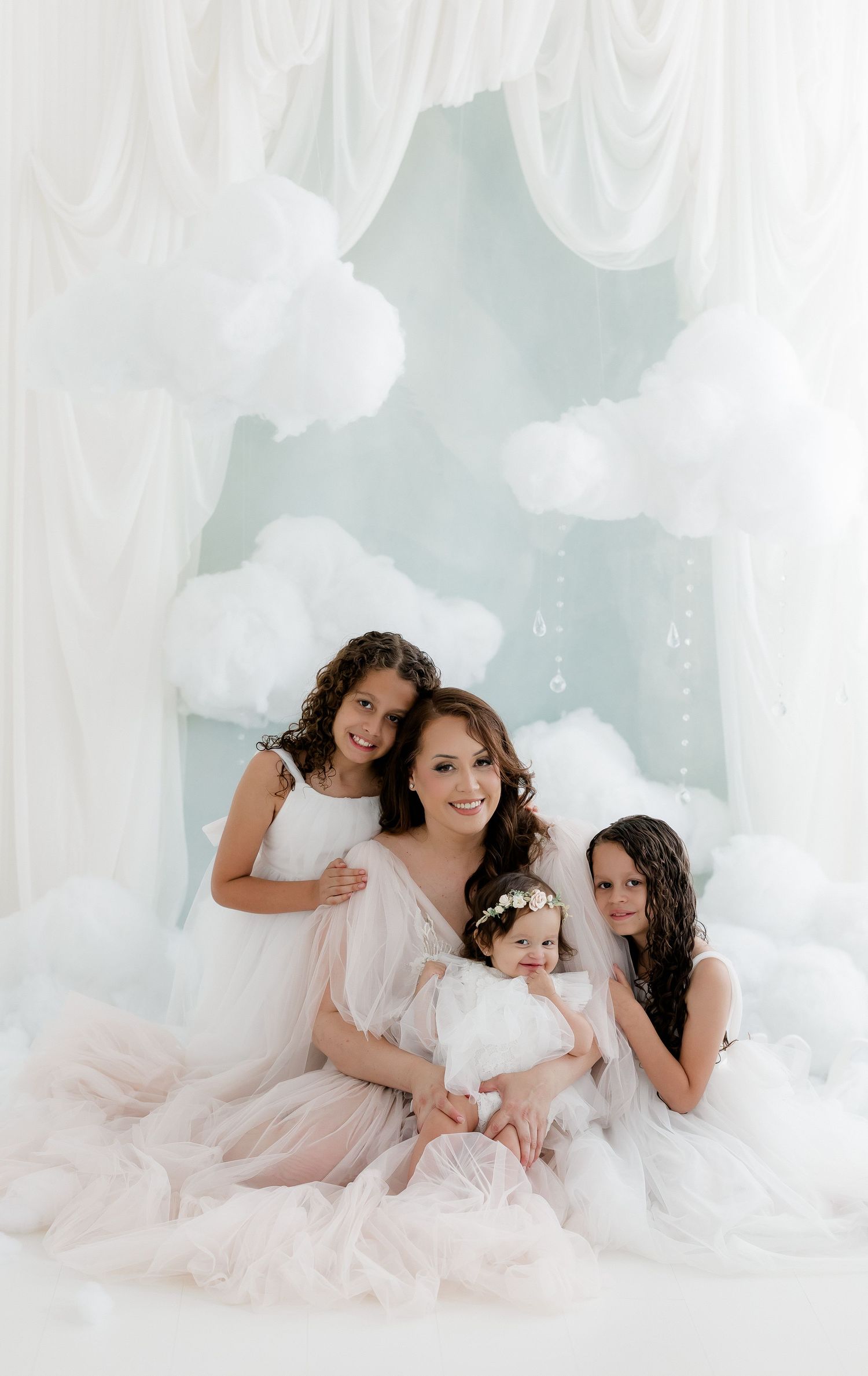 Pregnant mom poses with three daughters in white dresses surrounded by fluffy cloud decorations.