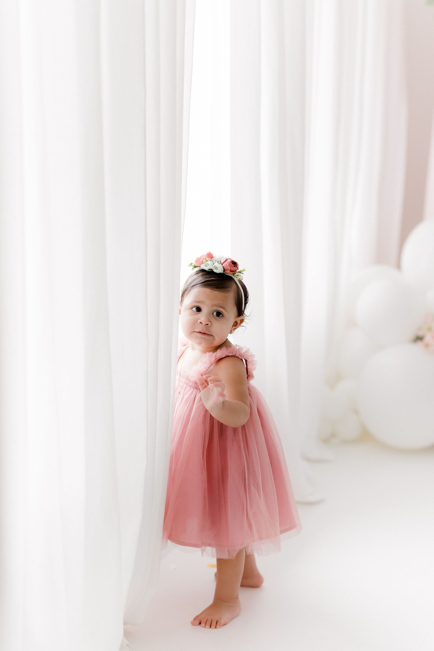 Baby in pink dress posing between white curtains in studio setting.
