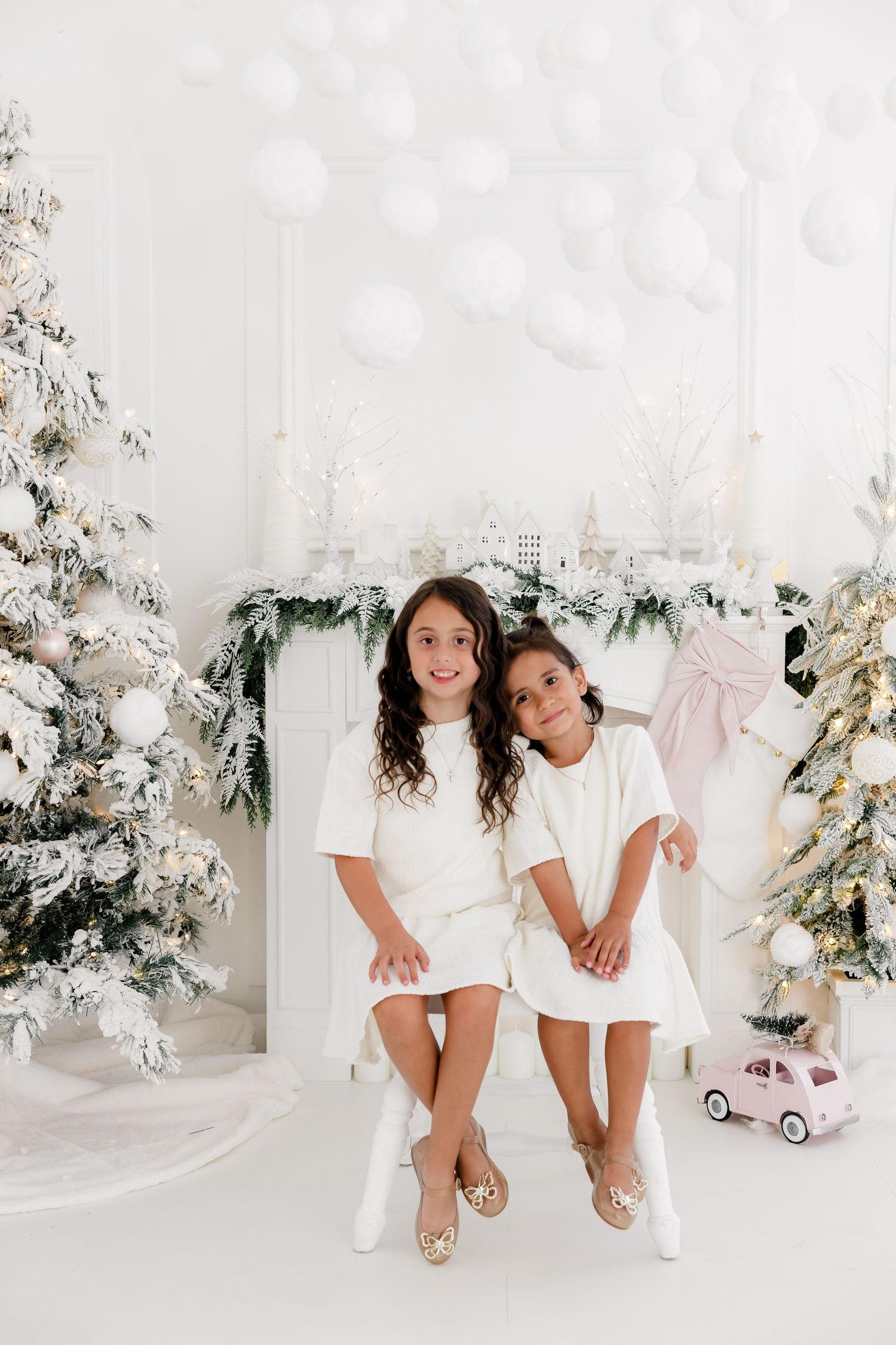 Two children in white dresses sit on a bench surrounded by snow-covered Christmas trees and decorations.