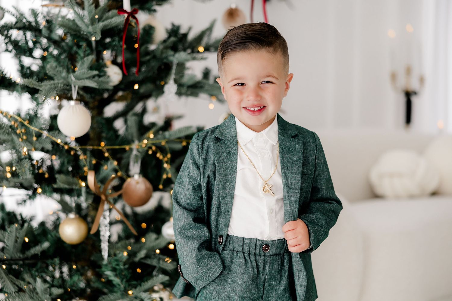 Young child in formal wear poses next to decorated Christmas tree in bright white studio setting.