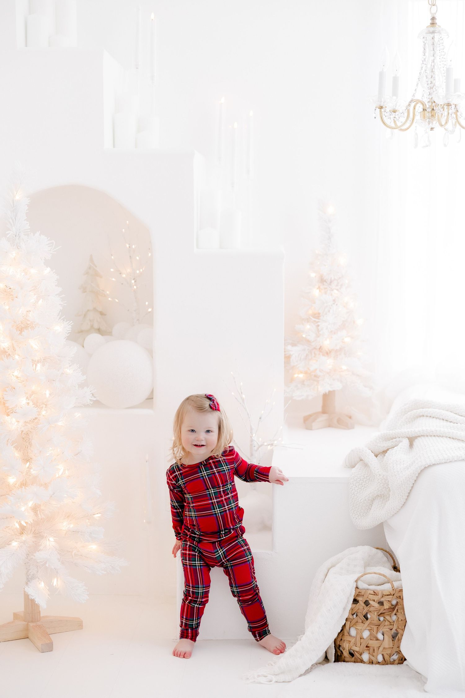 Baby in red plaid pajamas playing near white Christmas decorations and twinkling lights.