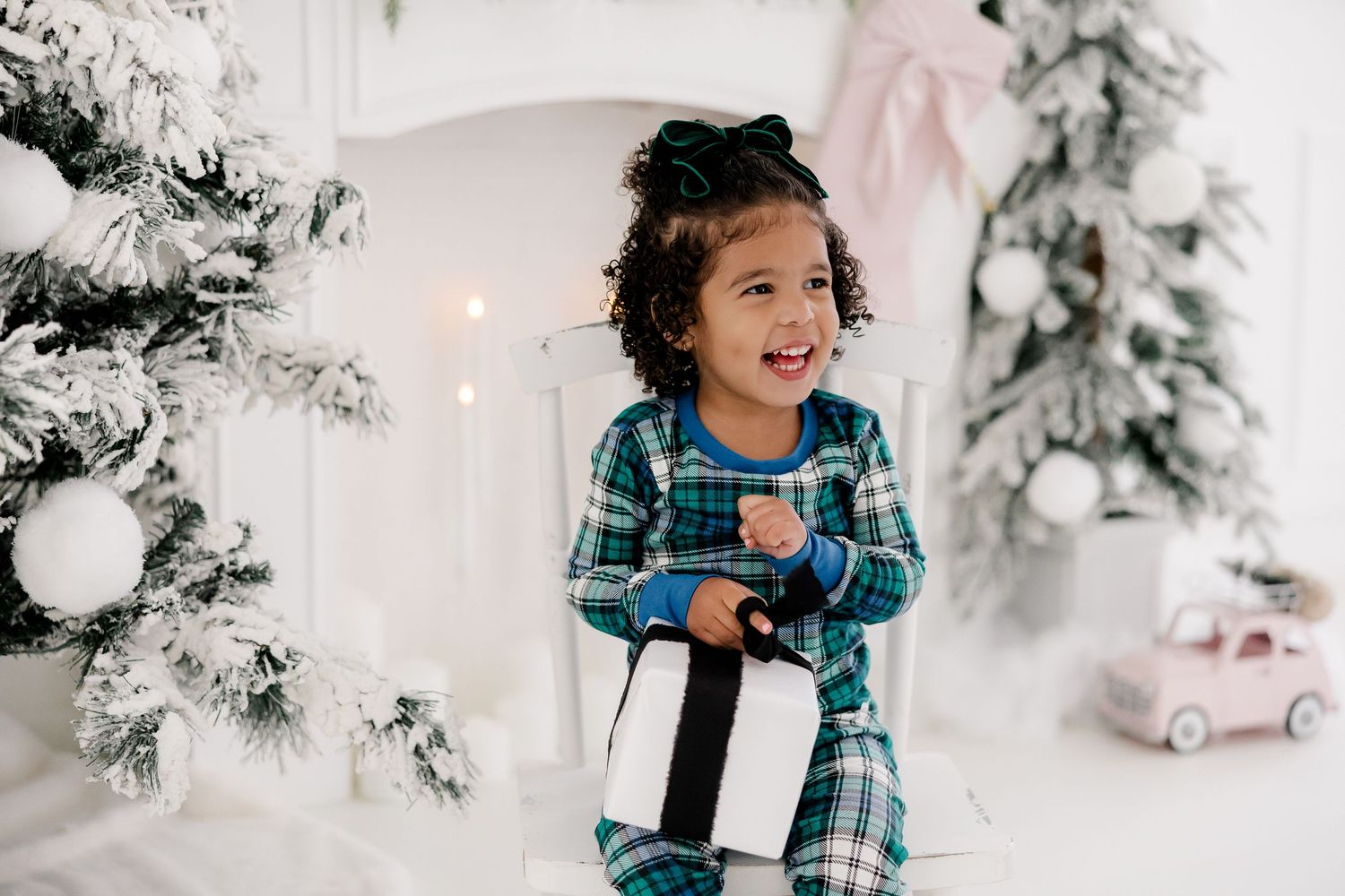 Child in blue plaid shirt plays joyfully in a bright white Christmas-themed studio setting with snow-covered trees.