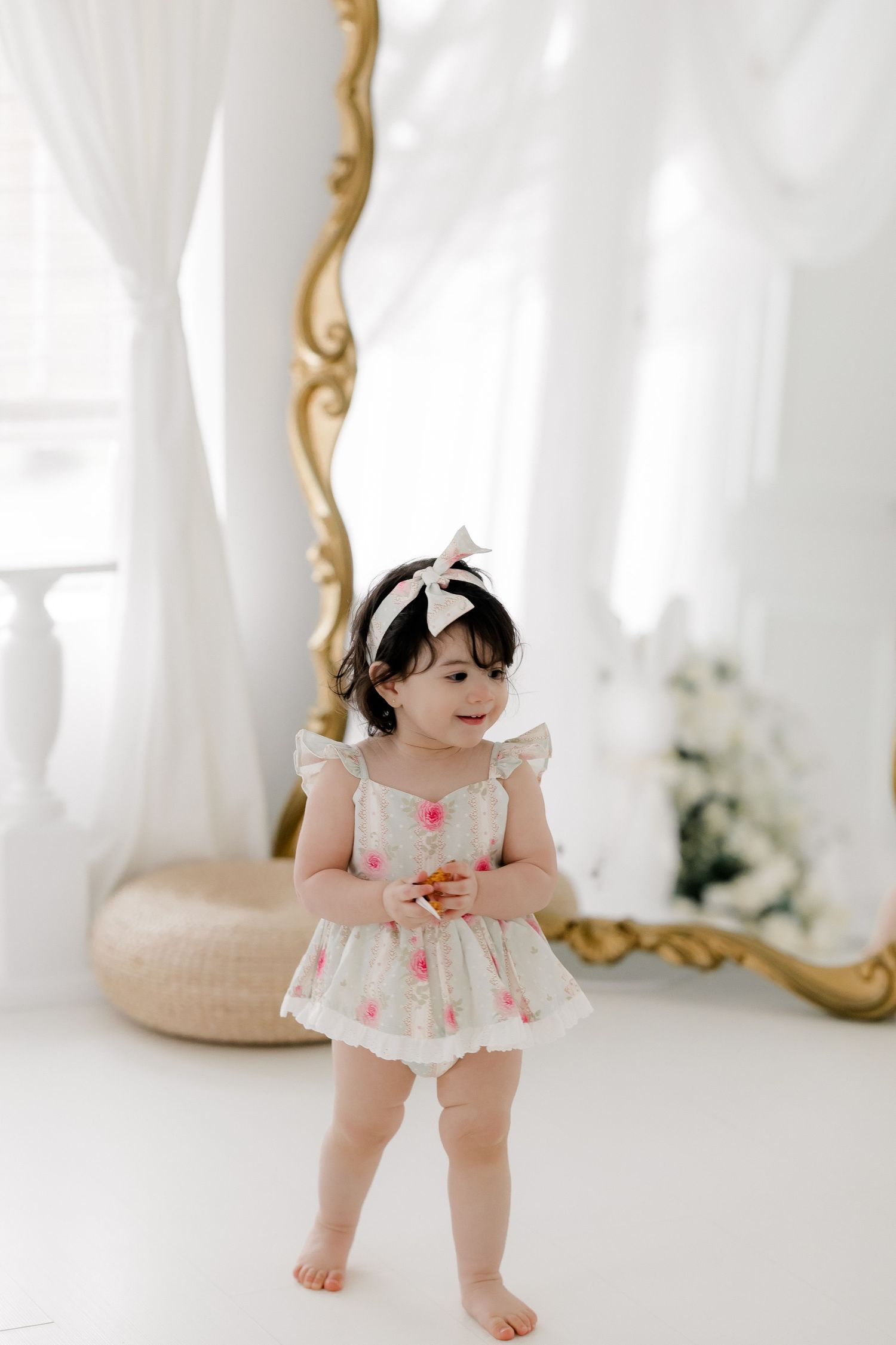 Baby wearing white ruffled dress and bow headband poses in bright studio setting with gold mirror frame.