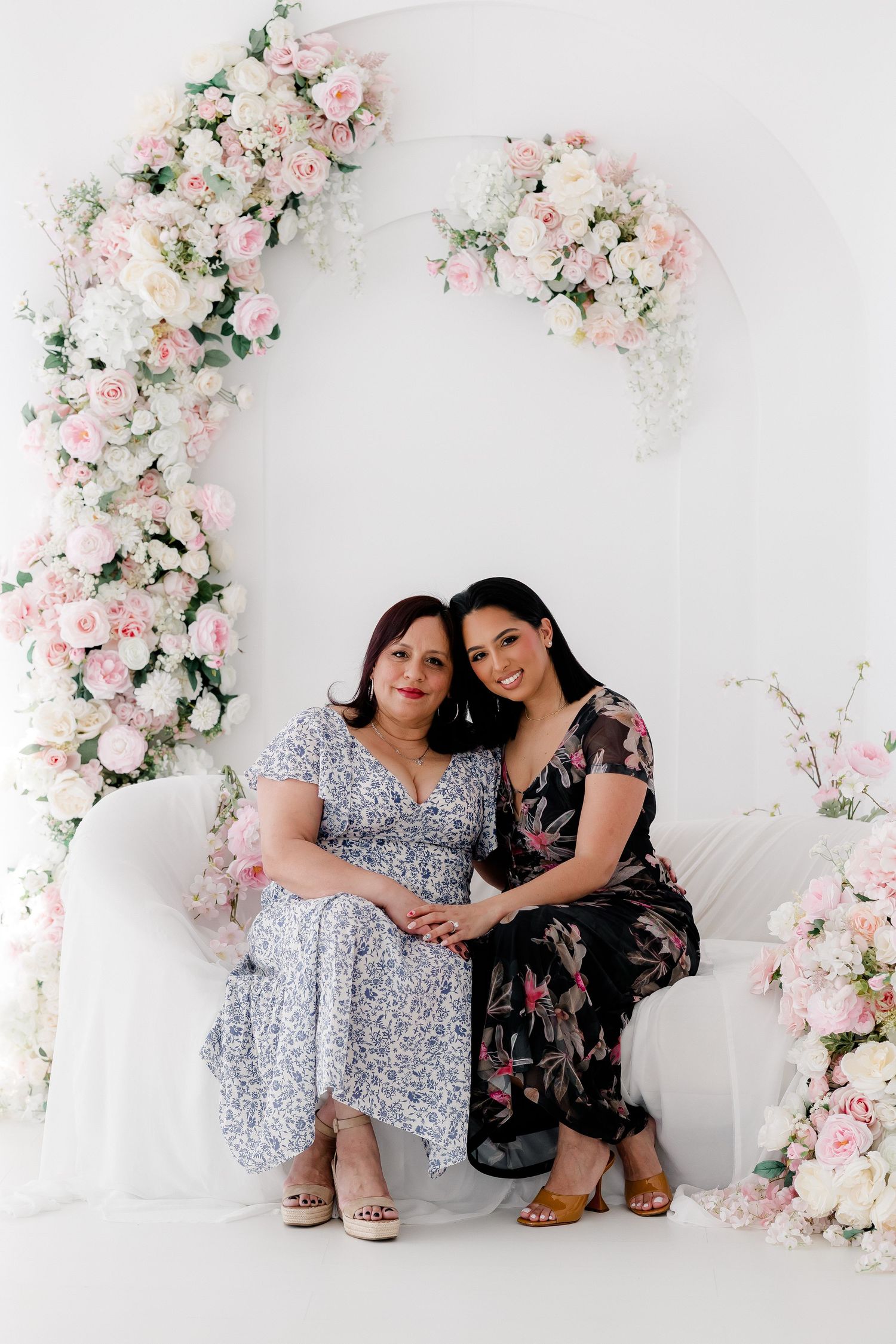 Two people pose together against a white wall decorated with delicate pink floral arrangements and greenery.