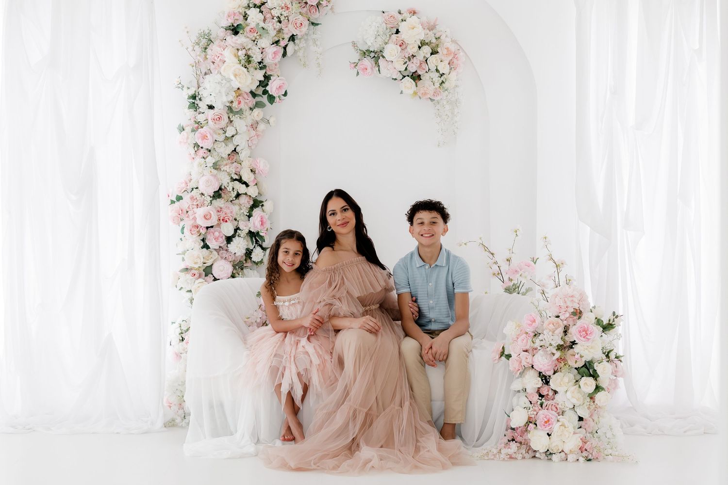 Elegant studio portrait with pink floral arch and white curtain backdrop in soft romantic lighting.