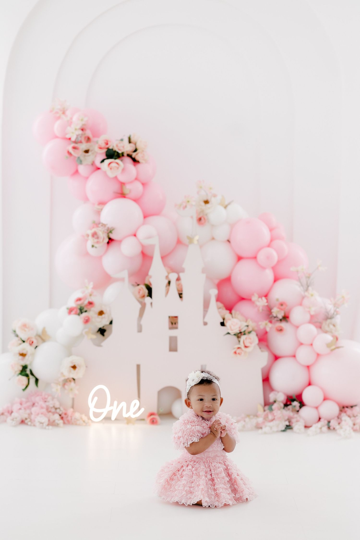 Pink and white balloon garland with floral accents creates a dreamy first birthday photo backdrop setup.