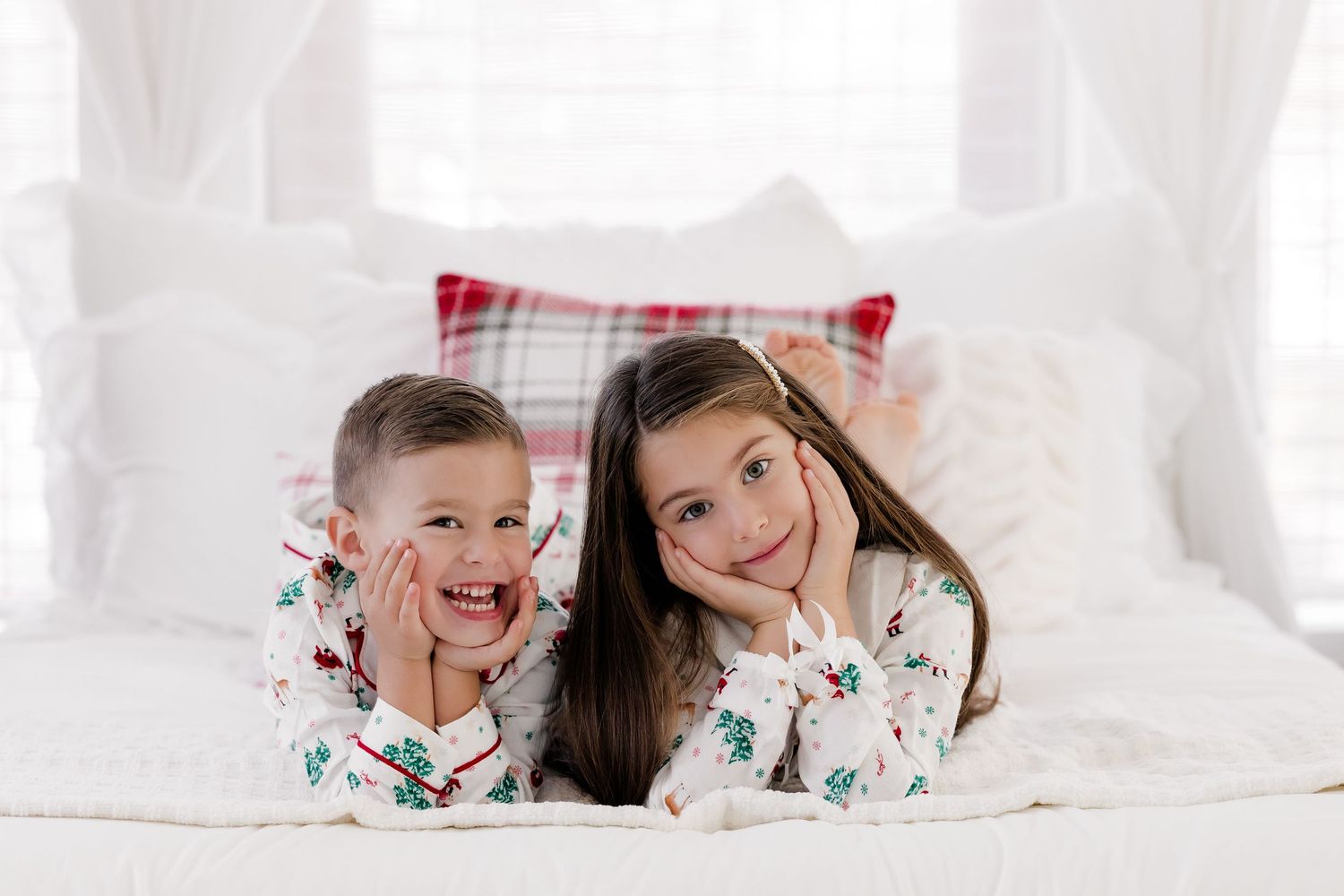 Two children in matching Christmas pajamas lying on a white bed, smiling with hands on cheeks.
