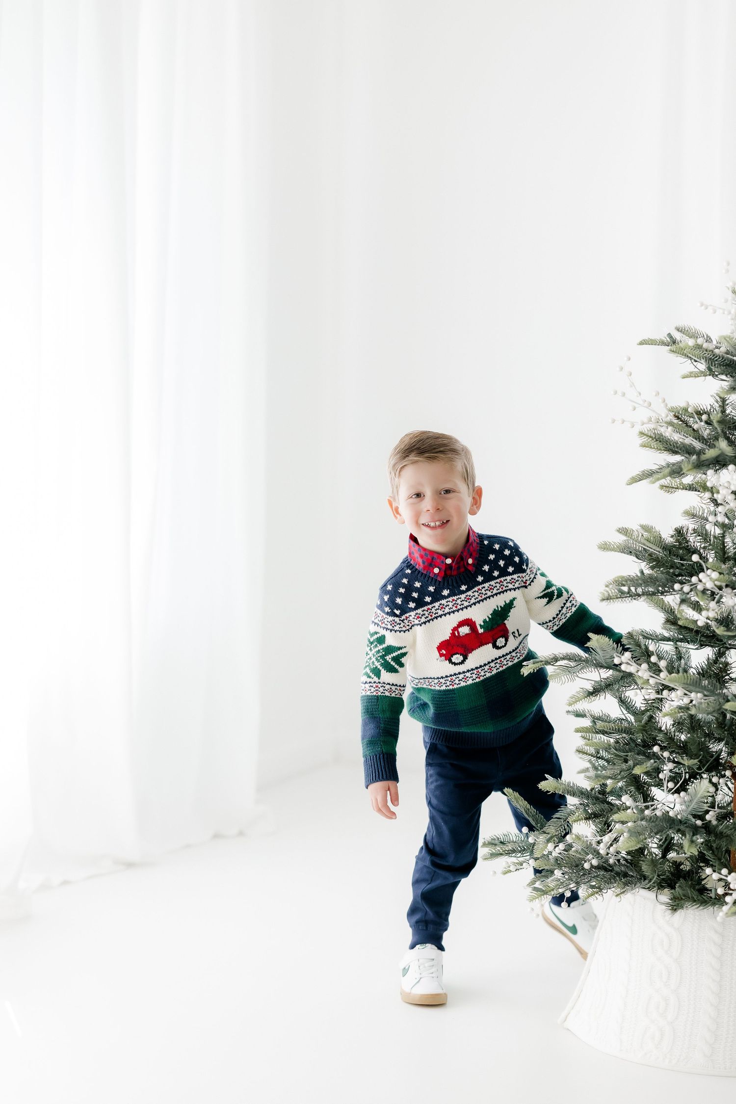A smiling boy in a festive Christmas sweater stands next to a decorated Christmas tree in a bright white room.