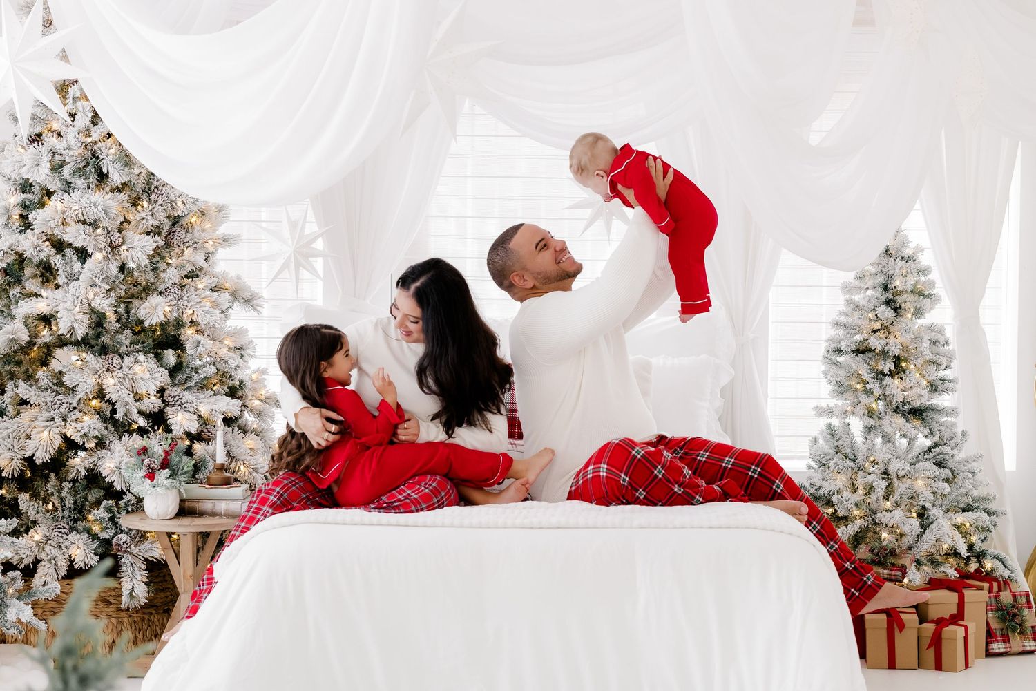 Family in red and white pajamas celebrating Christmas on a bed surrounded by decorated trees and gifts.