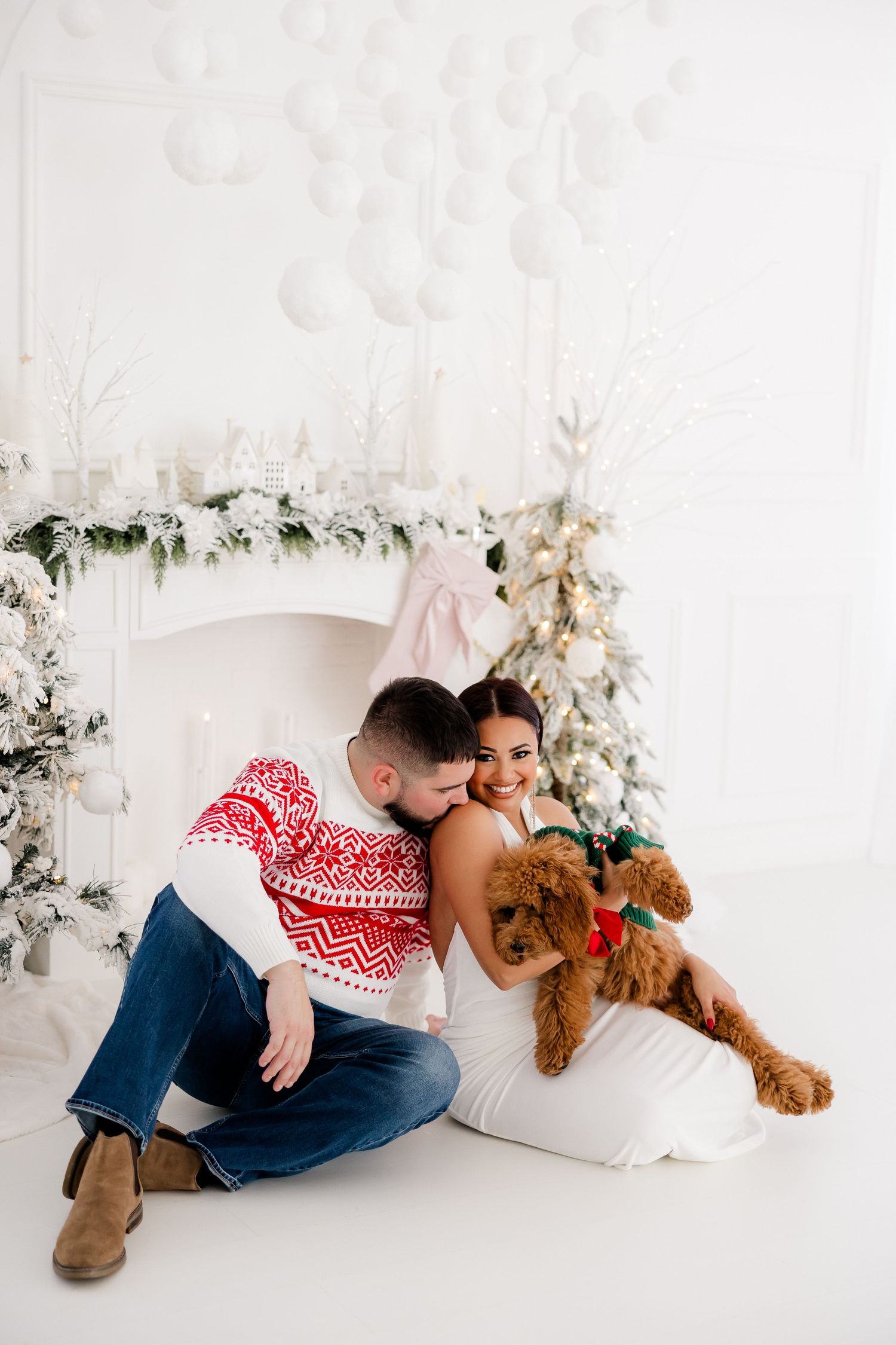 Couple cuddling with teddy bear in front of white Christmas tree and fireplace.