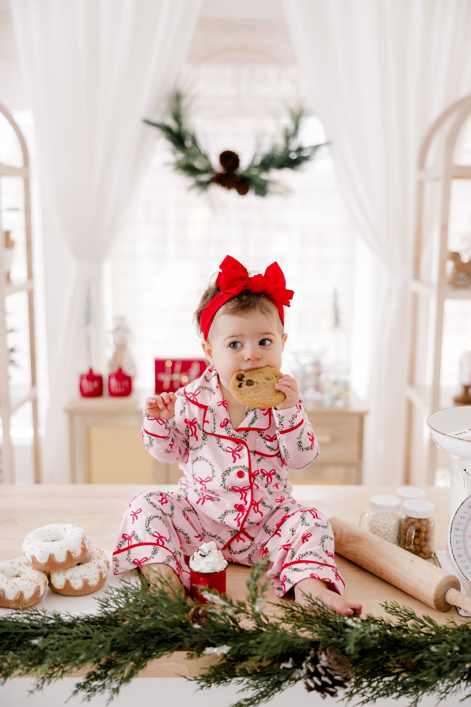 Baby in red bow and holiday pajamas eating cookie on decorated Christmas table setting.nj holiday mini sessions