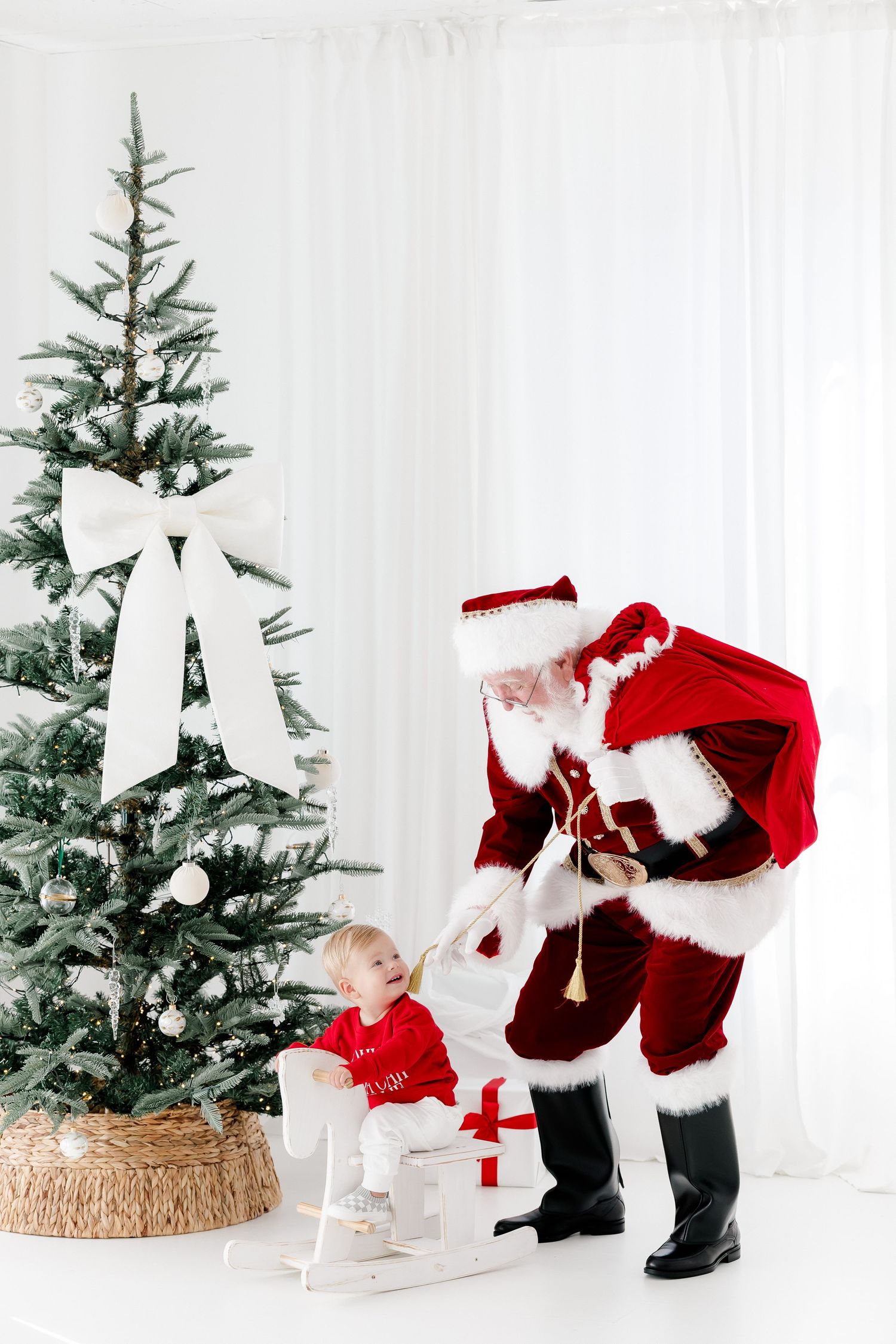 Santa Claus bending down to give a gift to a smiling toddler in red beside a decorated Christmas tree.
