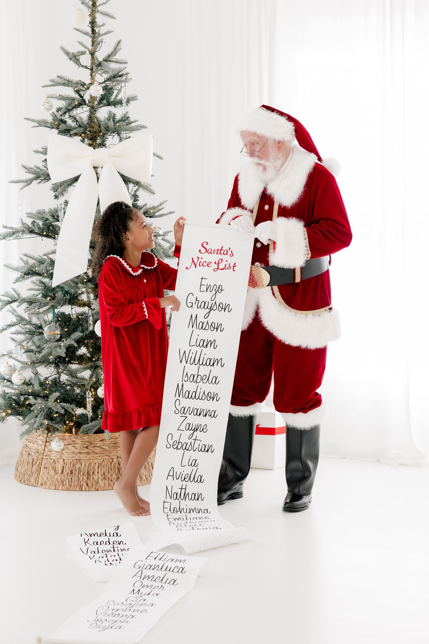A young girl in red dress reviews Santa's Nice List scroll with Santa Claus beside a decorated Christmas tree.